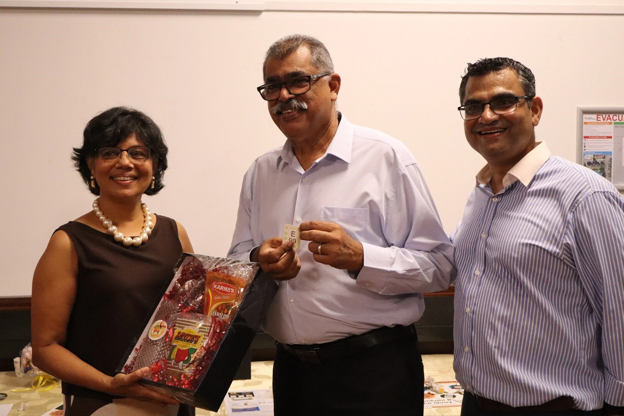 Three people smiling and standing indoors, with one holding a gift and another holding a small card.