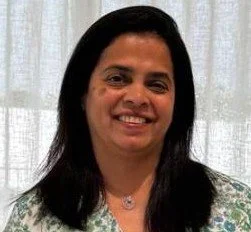 Portrait of a smiling woman with long dark hair, wearing a necklace, standing in front of a light-colored curtain.