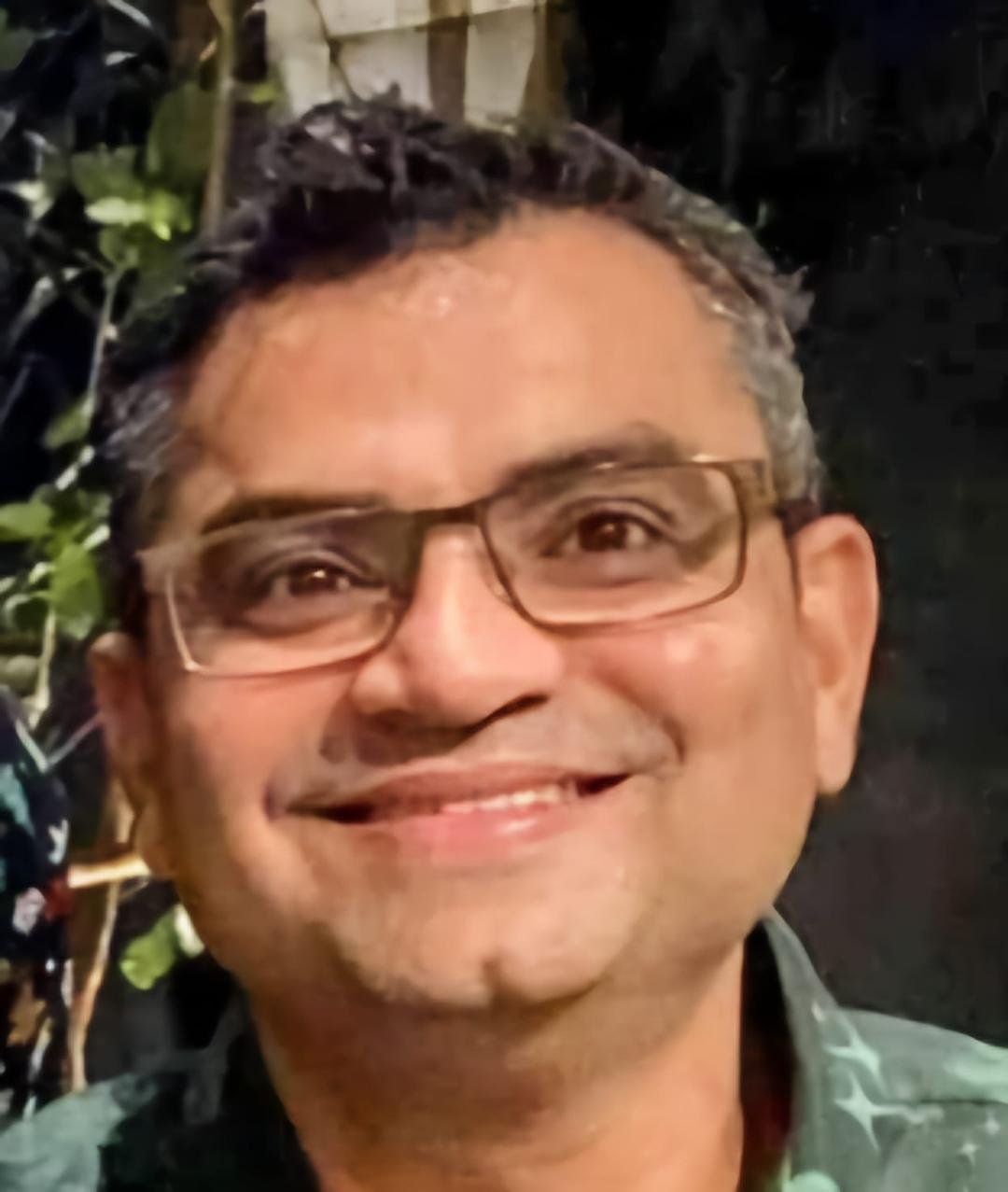 Close-up of a man smiling, wearing glasses, with dark hair, in an outdoor setting with greenery in the background.