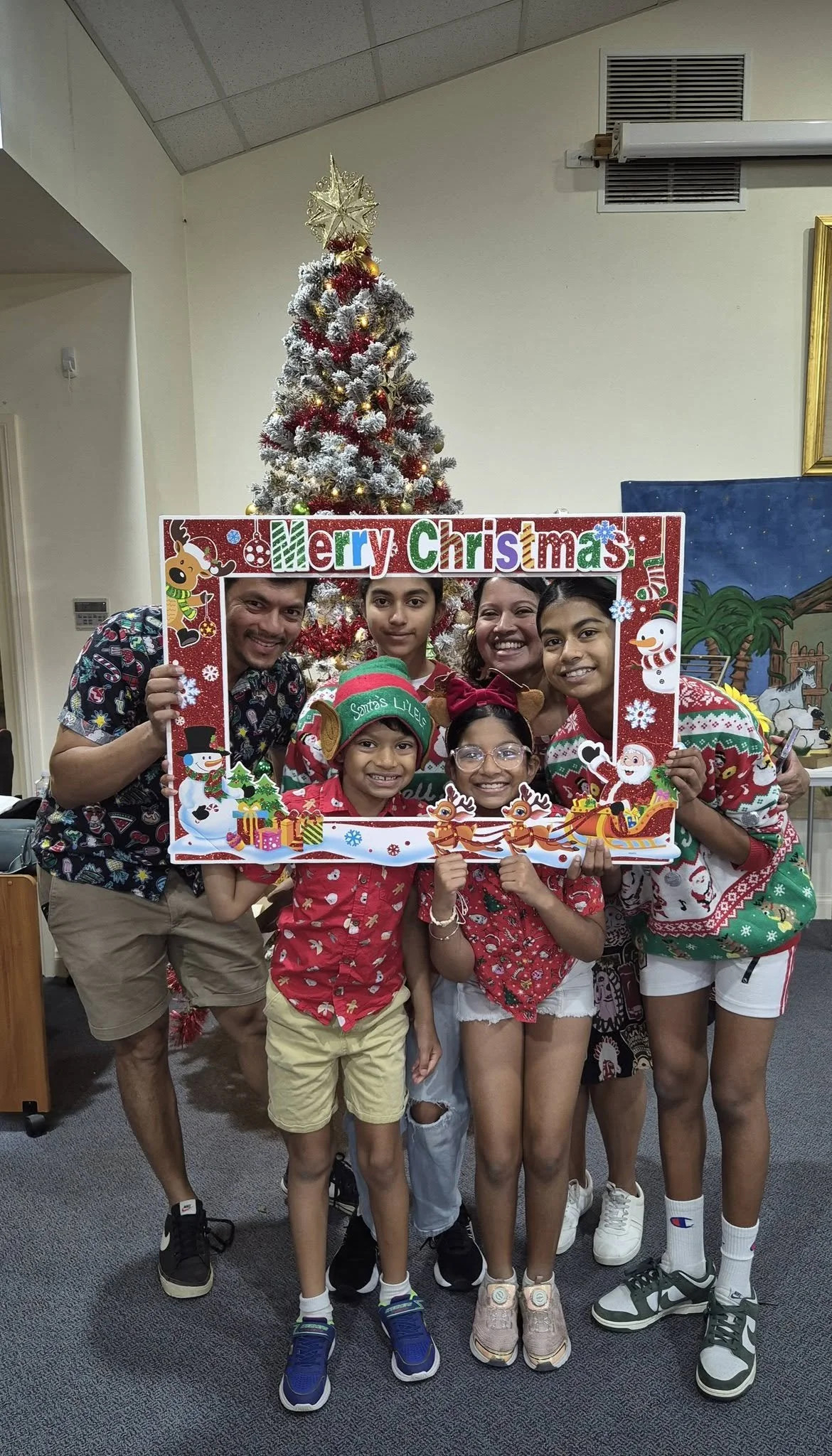 A group of six people, including children and adults, smiling and standing behind a festive frame with a Christmas theme, in front of a decorated Christmas tree. They are indoors, celebrating Christmas.