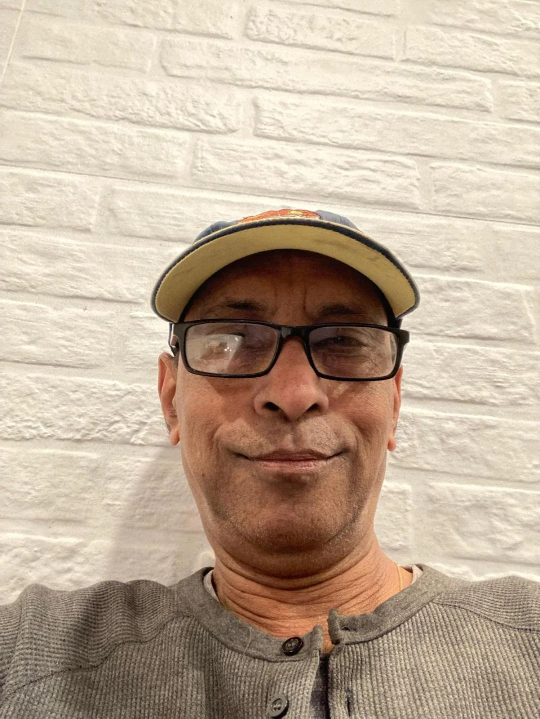 Close-up of a middle-aged man wearing glasses, a baseball cap, and a gray sweater, smiling in front of a white brick wall.