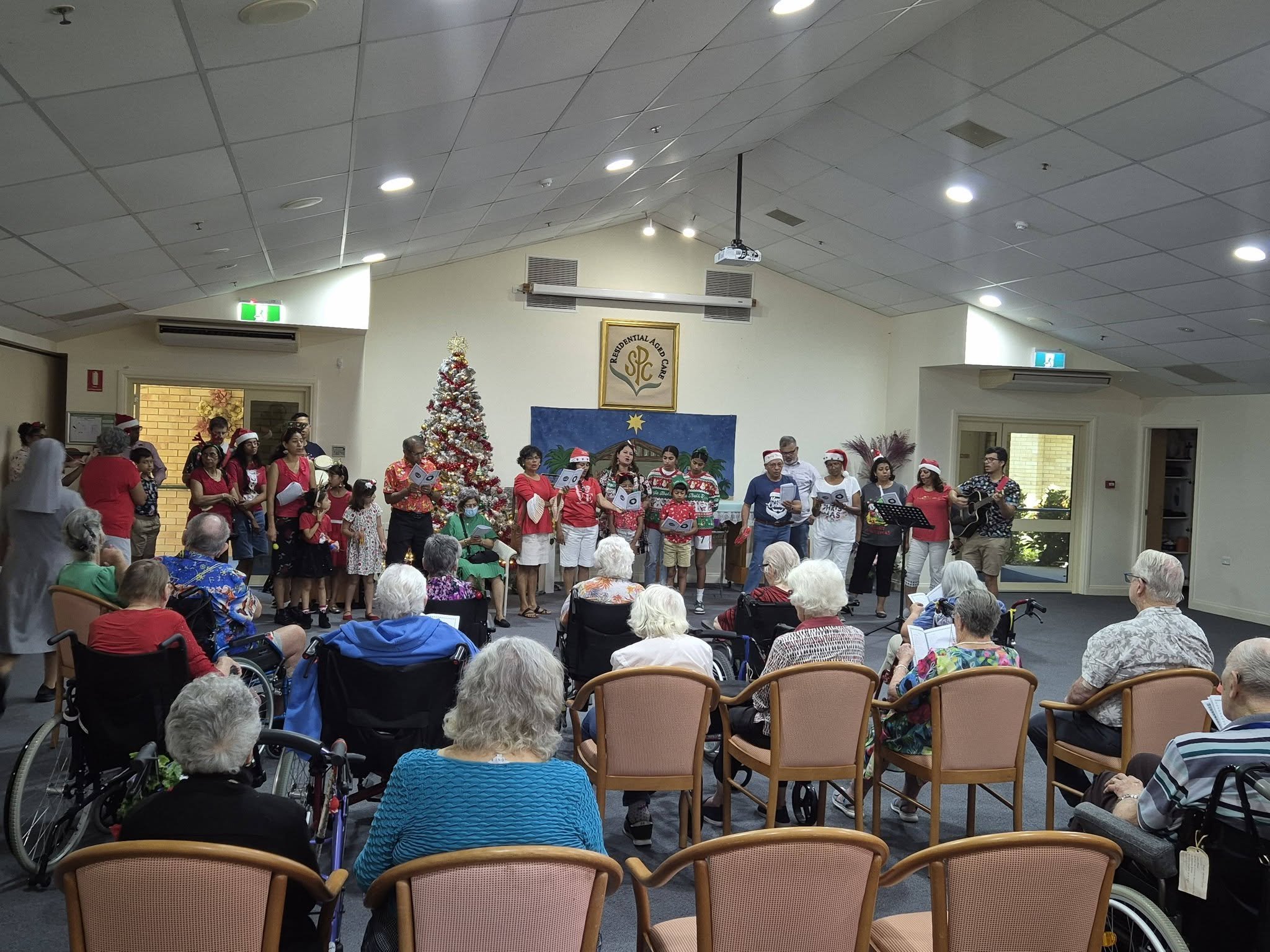 A Christmas choir performing in front of an audience of elderly residents, with a decorated Christmas tree, at what appears to be a senior care facility.