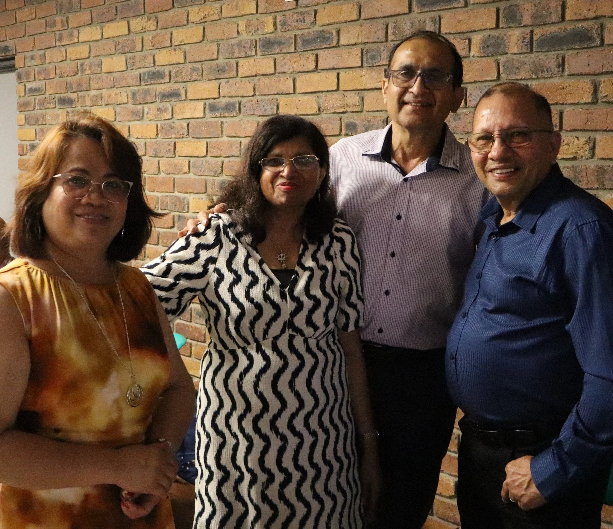 Four adults standing close together in front of a brick wall, smiling at the camera. Two women and two men, dressed in semi-formal attire.