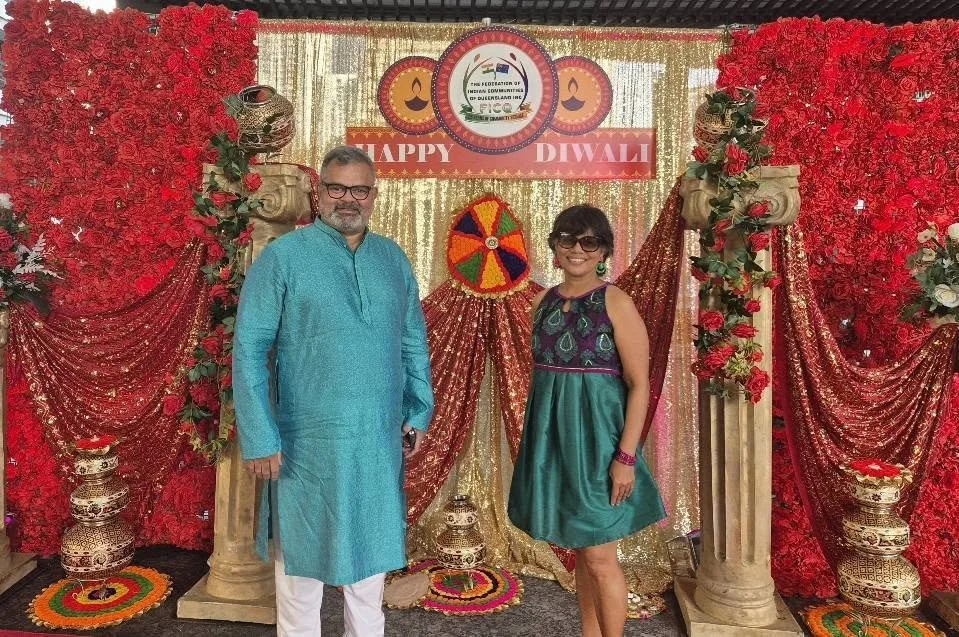 A man and woman standing in front of a decorated backdrop with red flowers, gold curtains, and a sign that reads 'Happy Diwali'. The backdrop features traditional Indian elements such as ornate pillars and colorful decorations, indicating a celebrati