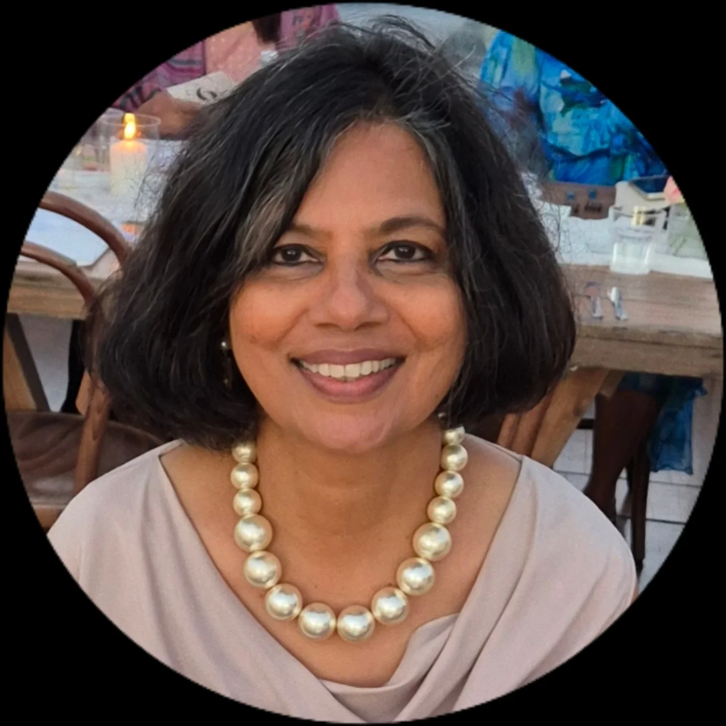 Portrait of a smiling woman with shoulder-length black hair wearing a pearl necklace and a beige top, seated indoors at a dining area with lit candles and wood tables.