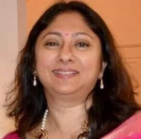 A woman with long dark hair wearing a pink outfit and pearl jewelry, smiling at the camera.