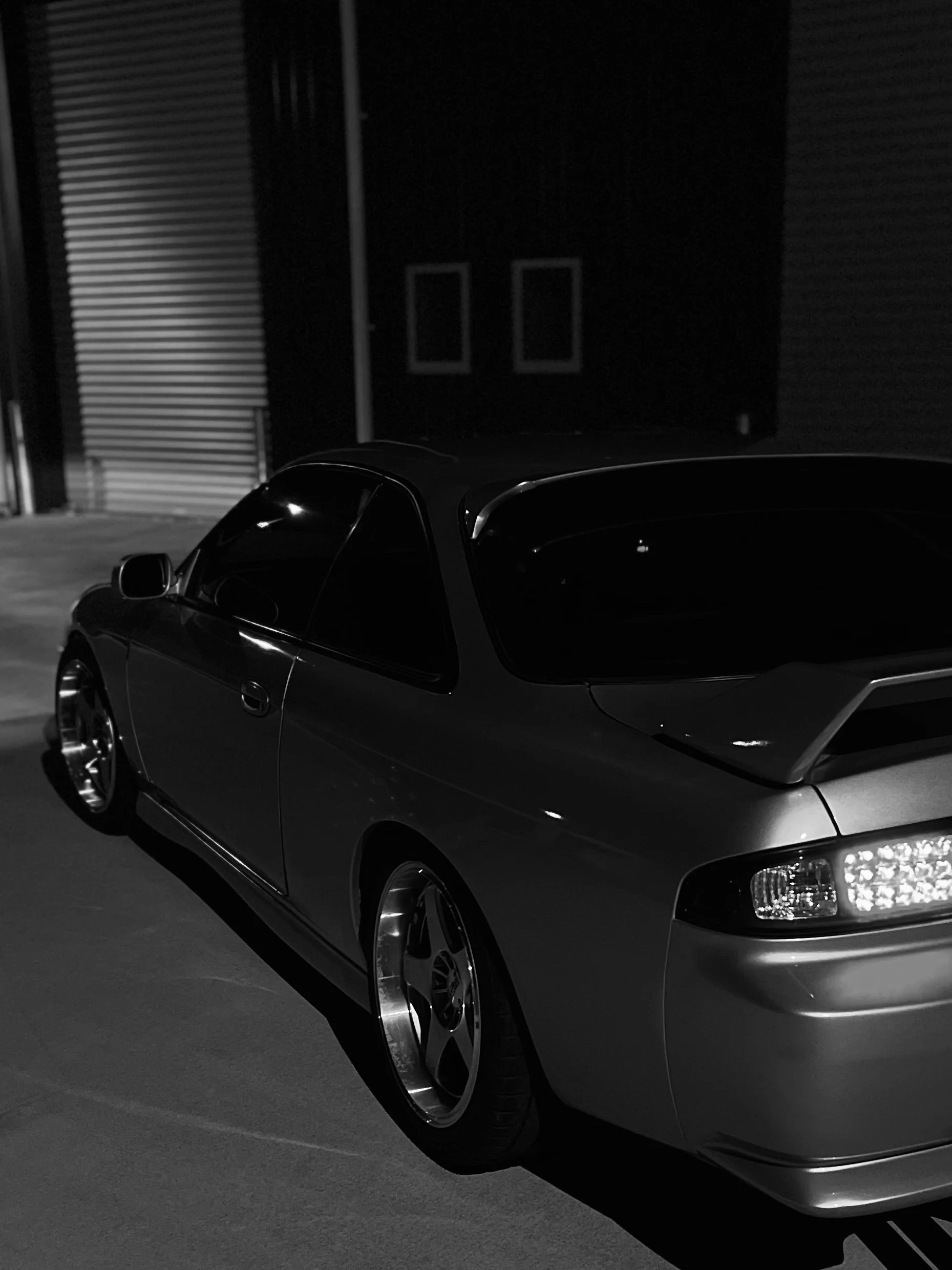 A dark-colored sports car parked in front of industrial building at night. The car has sleek design, tinted windows, and shiny rims.