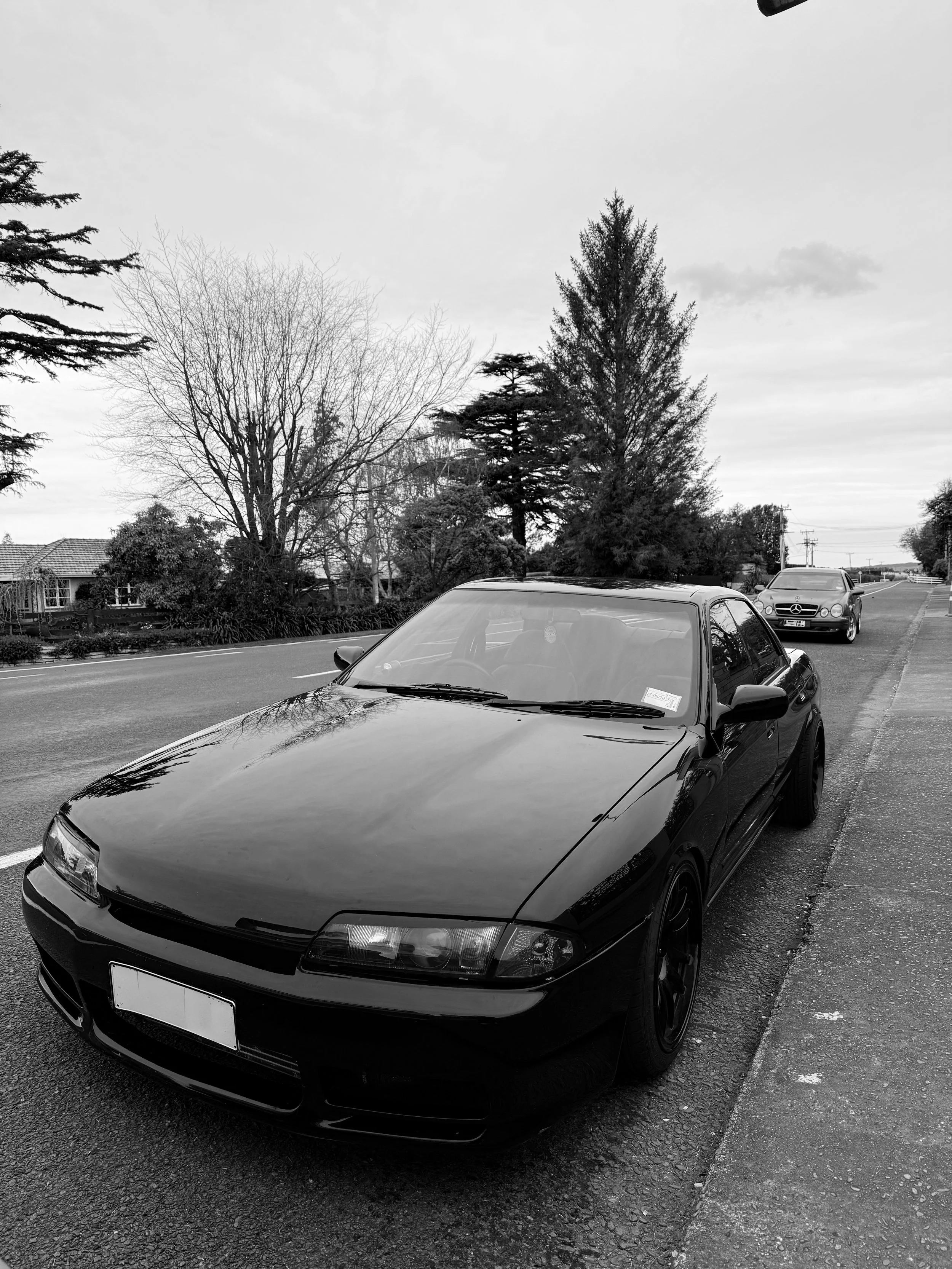 Black sports car parked on the side of the street with trees and another car in the background, in a suburban neighborhood, in black and white.