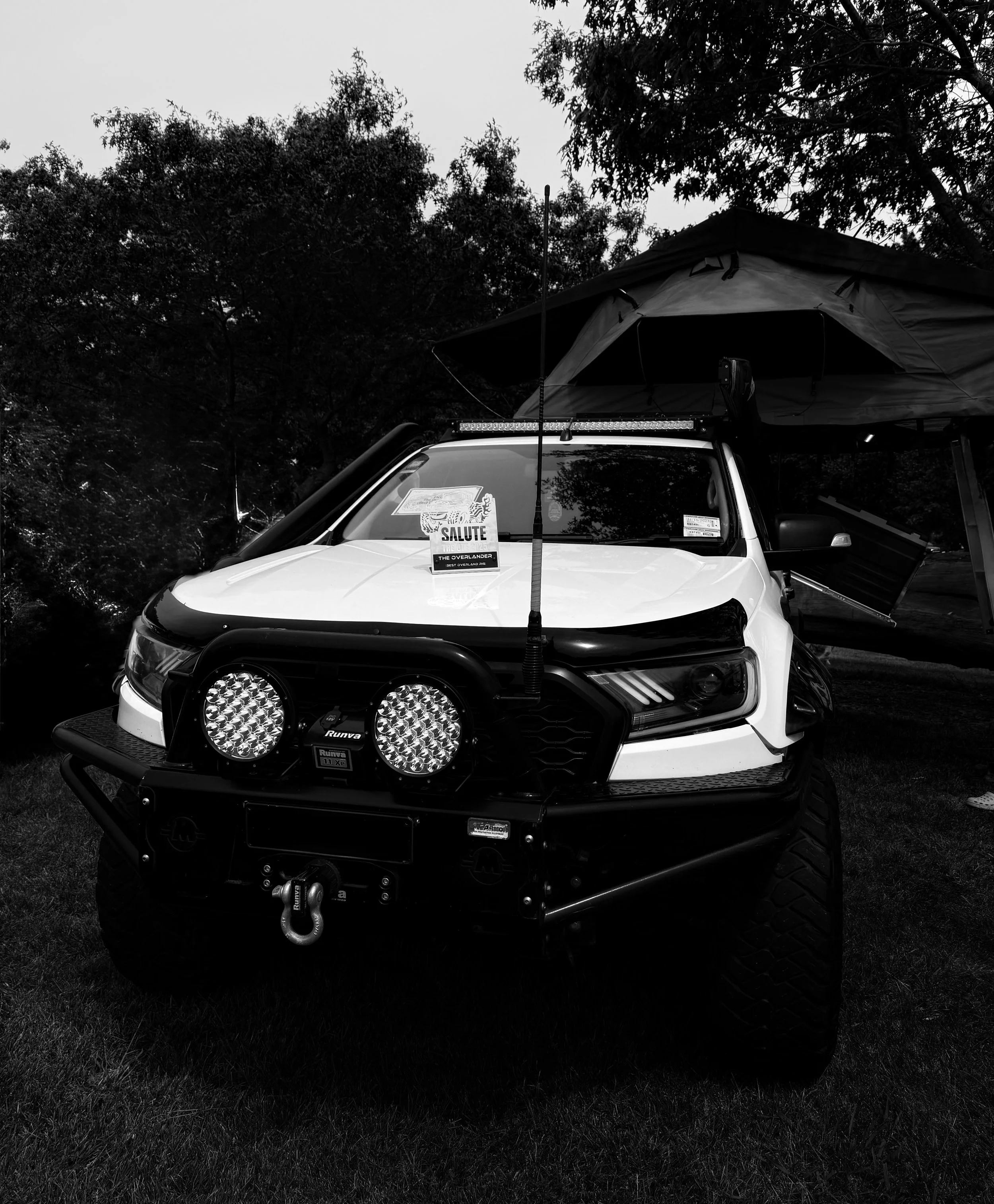 A white off-road vehicle with a roof tent, large tires, and additional lighting on the front, parked on grass with trees in the background.