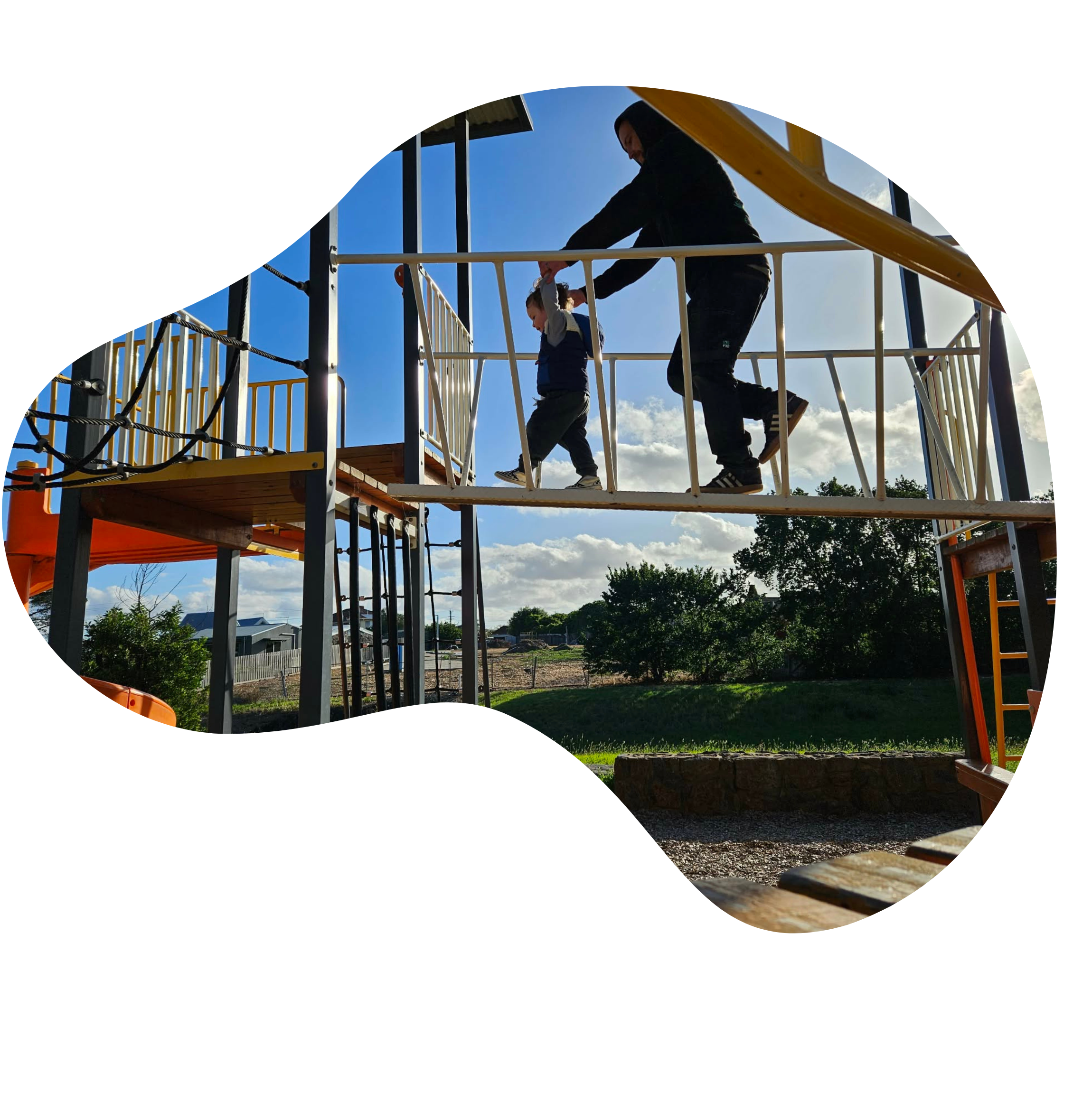 A child being helped by an adult to walk across a bridge on a playground structure, with a bright blue sky and green trees in the background.