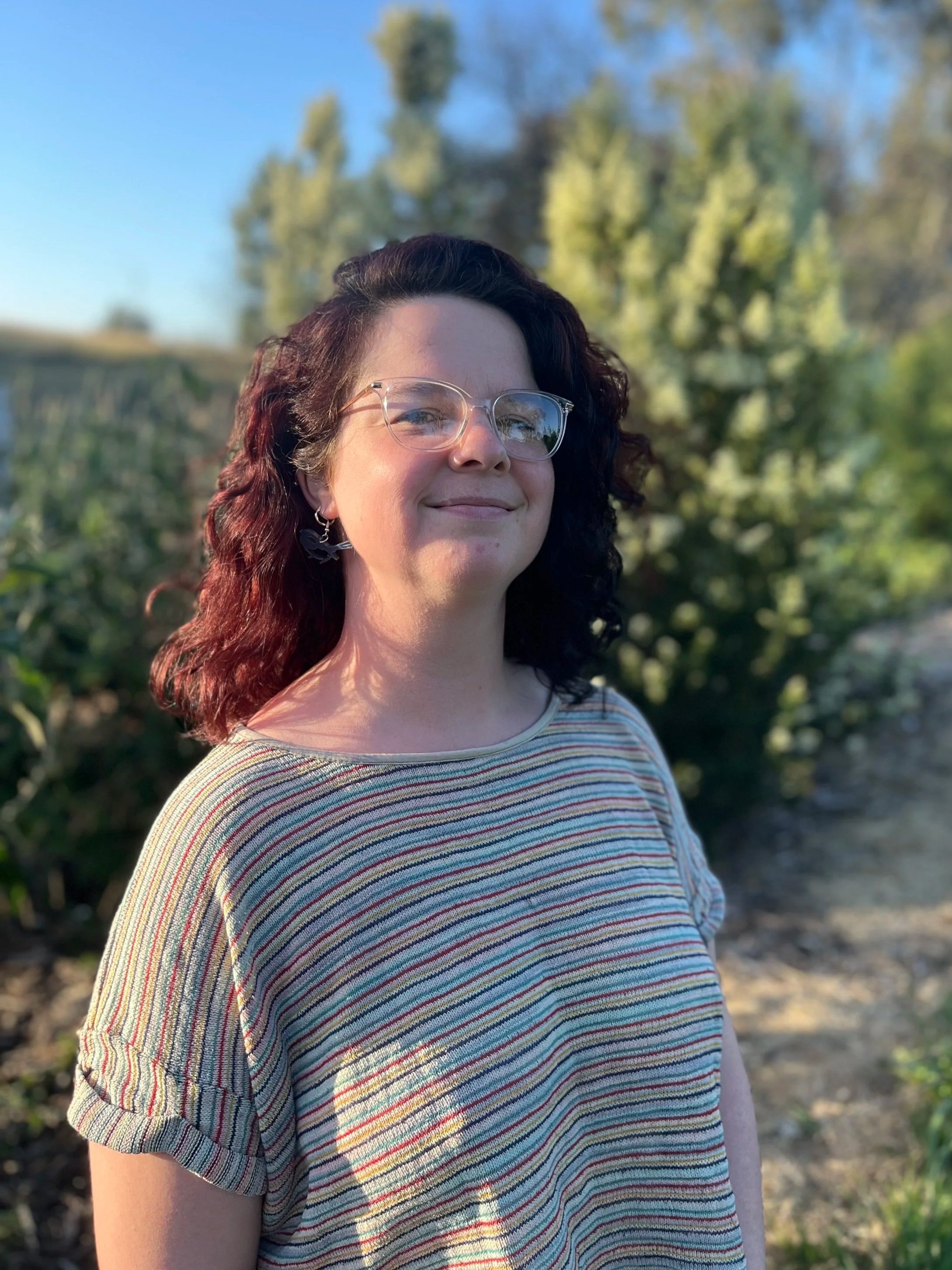 A woman, Monique Hale,  with curly dark hair and glasses smiling outdoors in front of green trees and bushes during daylight.