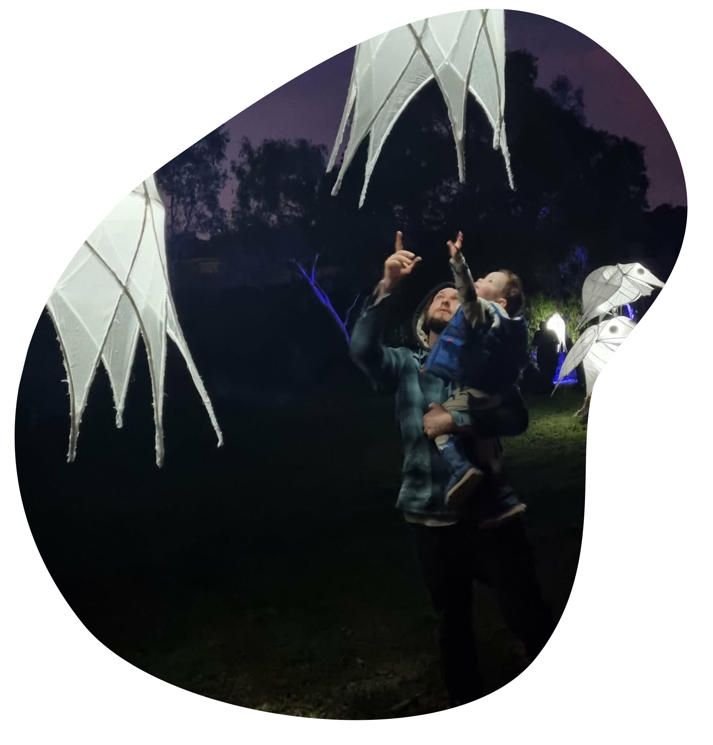 A man holding a young child while pointing at illuminated art installations resembling white, winged umbrellas or jellyfish, set outdoors at dusk or night with a dark sky and trees in the background.