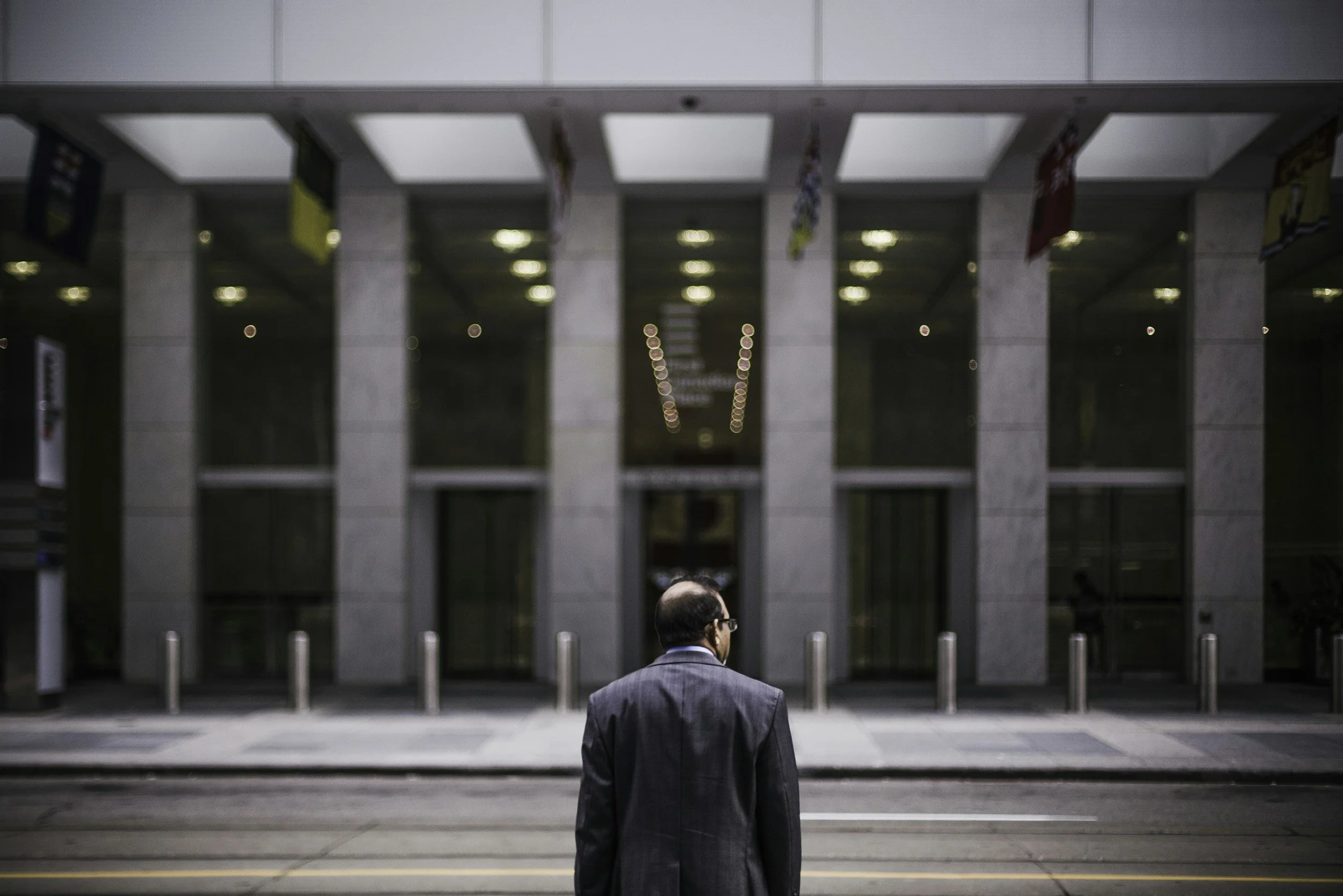 A man in a suit walking in front of a large modern building with glass windows, illuminated interior lights, and flags hanging from the ceiling inside.