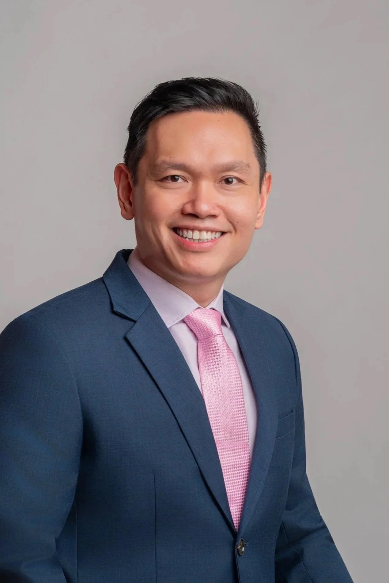 Portrait of a smiling man wearing a navy blue suit, pink tie, and white shirt, posed against a plain light gray background.