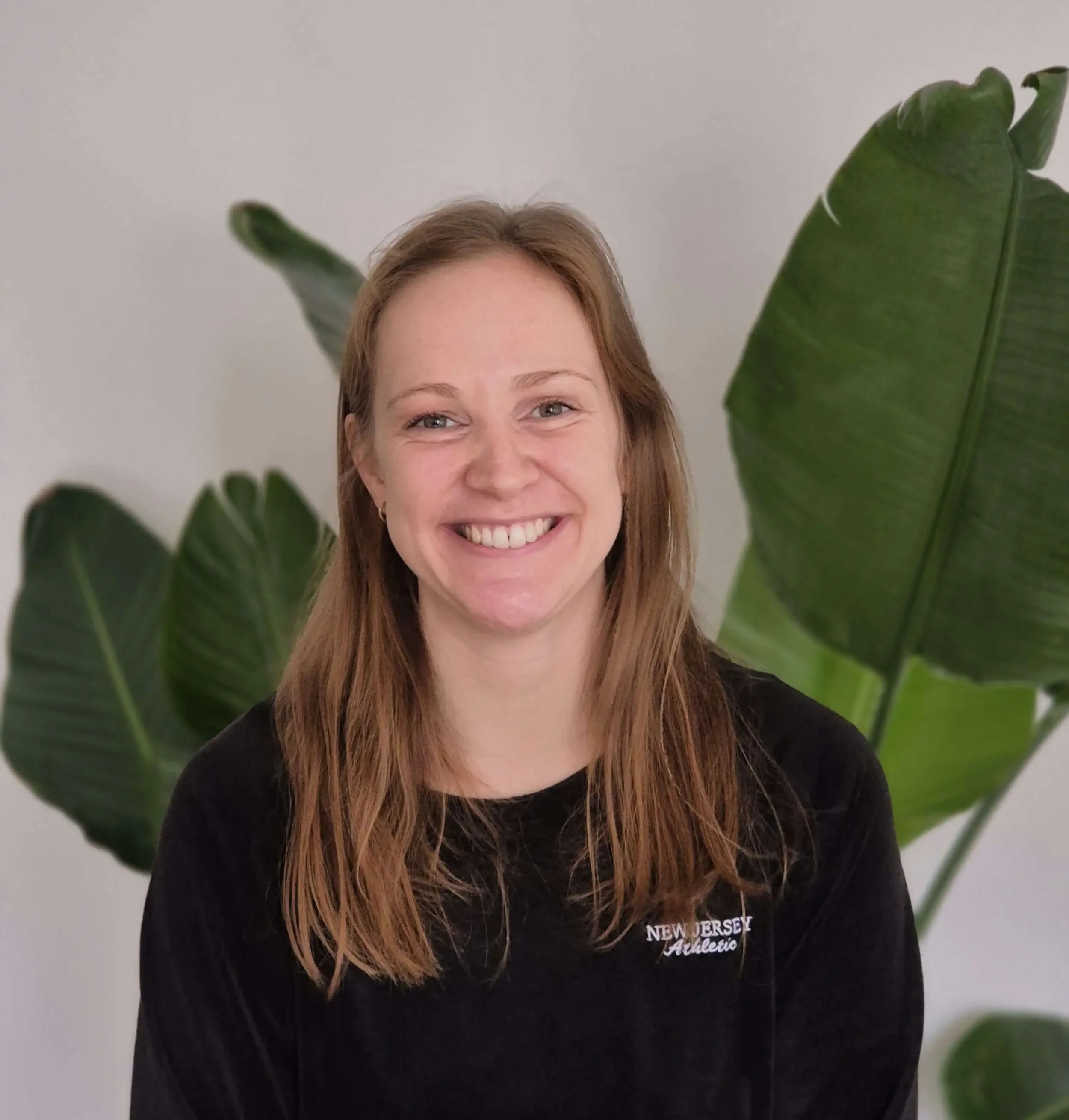 Lächelnde Frau mit braunen Haaren vor großen grünen Pflanzen an einer weißen Wand.