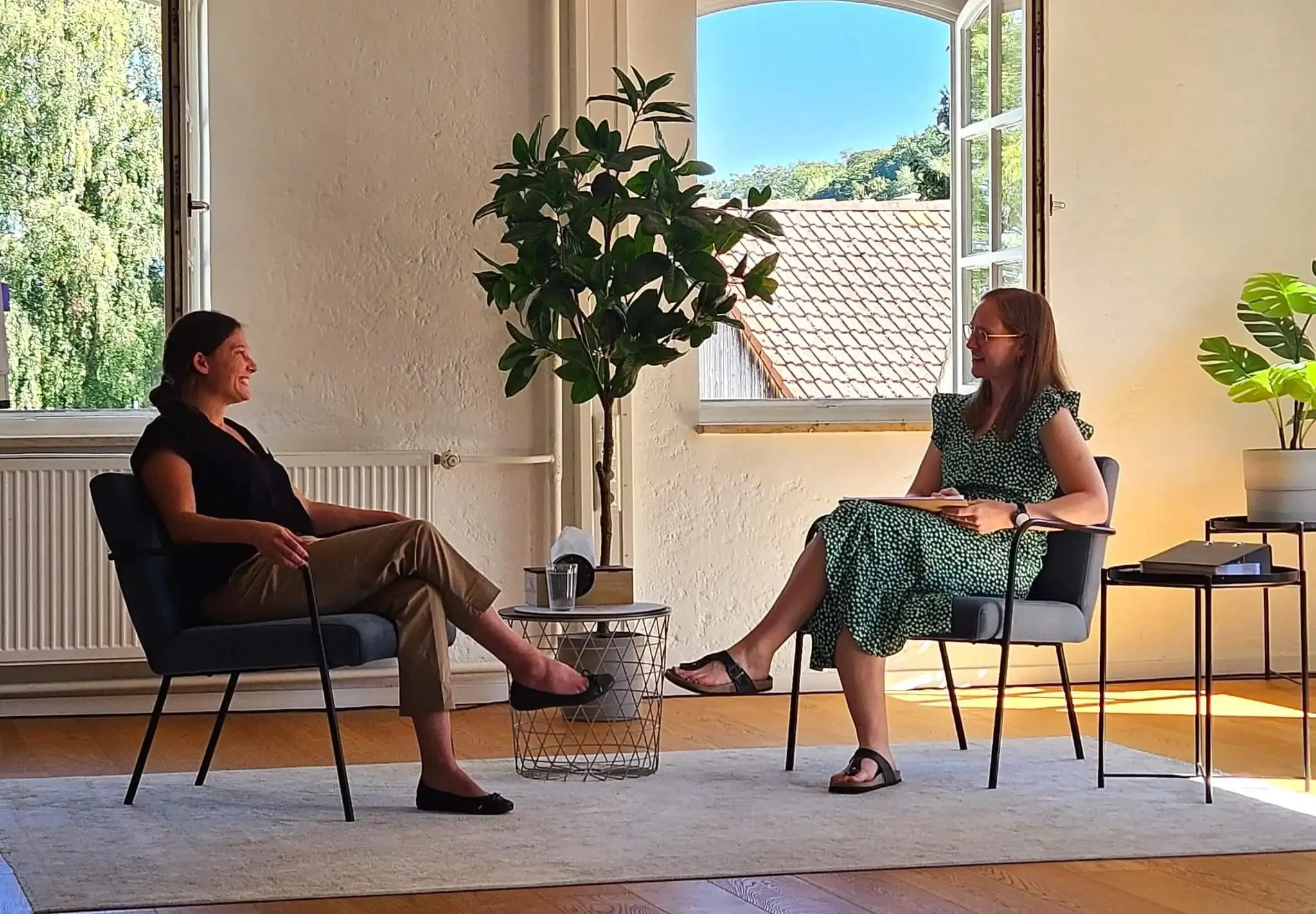 Zwei Frauen sitzen sich gegenüber in einem Raum mit großen offenen Fenstern, grünen Pflanzen und natürlichem Licht. Sie führen ein Gespräch, eine schreibt Notizen, beide lachen.