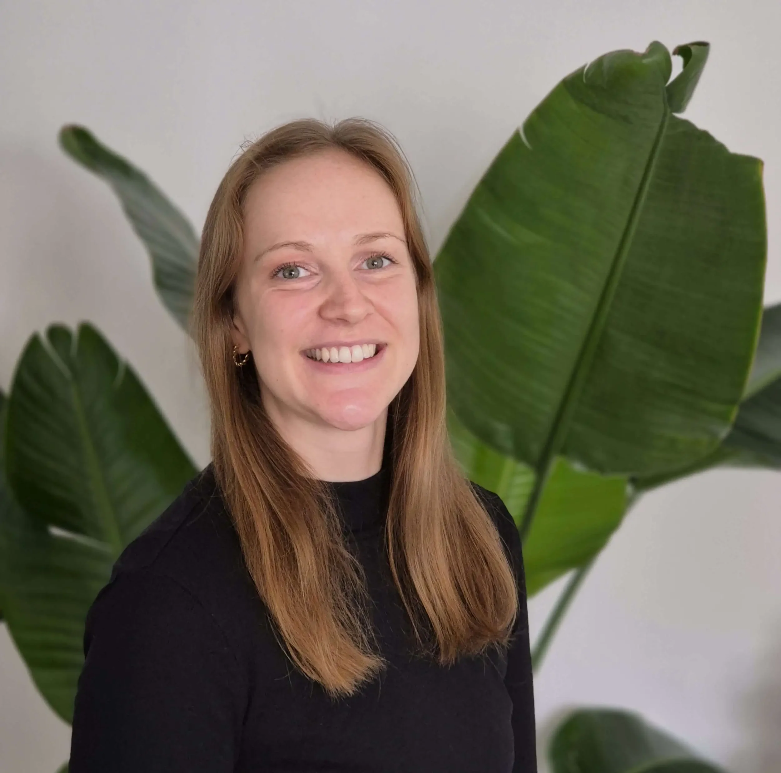 Lächelnde Frau mit langem, rotbraunem Haar trägt schwarze Kleidung vor einer grünen Pflanze mit großen Blättern.