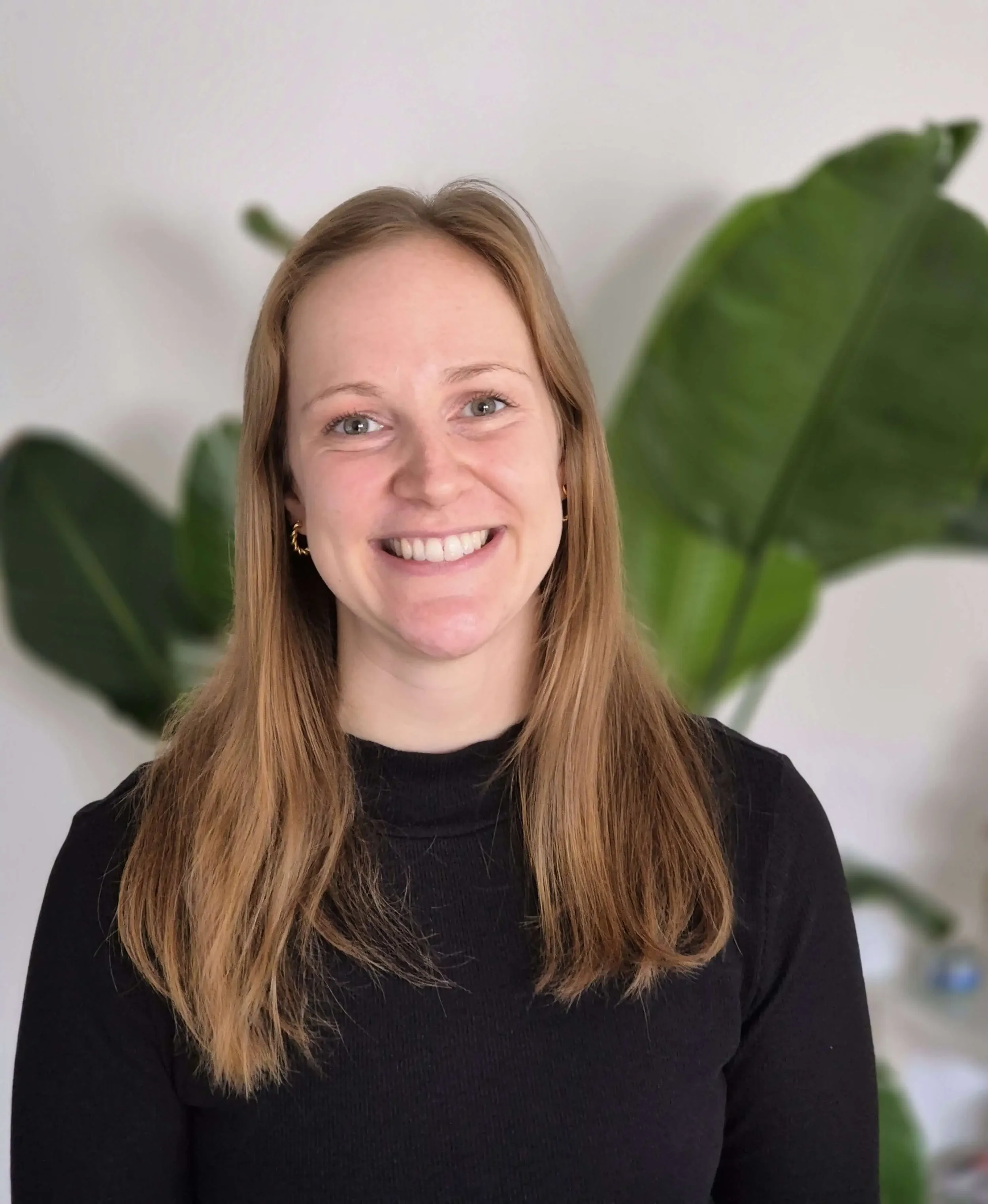 Ein Lächeln tragende Frau mit langen, braunen Haaren, dunklem Pullover und goldenen Ohrringen, vor grünen Blättern im Hintergrund.