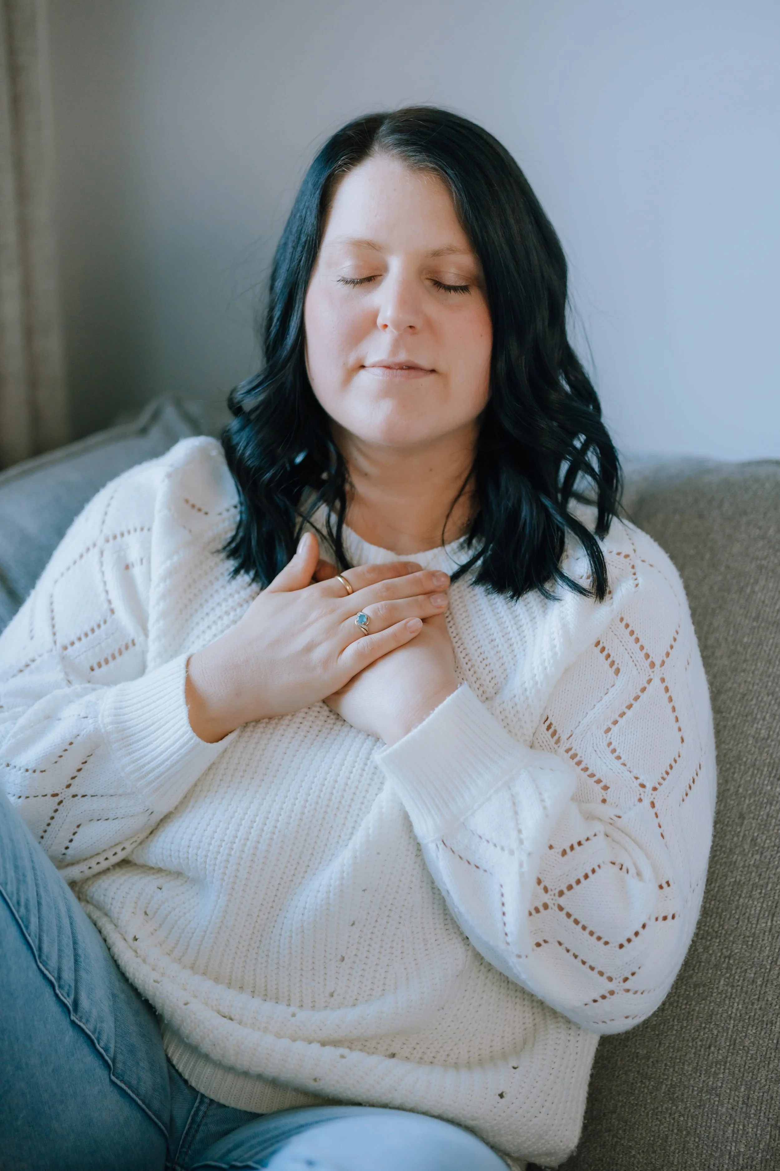 A woman with black hair and fair skin, sitting on a couch with her eyes closed and hands on her chest, wearing a white knitted sweater, appears to be meditating or practicing mindfulness.