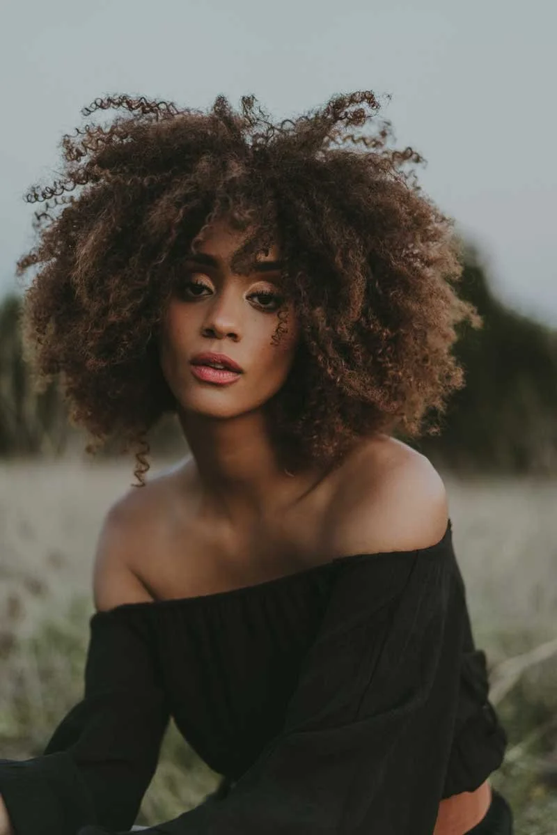 Young woman with curly brown hair wearing an off-the-shoulder black top outdoors.