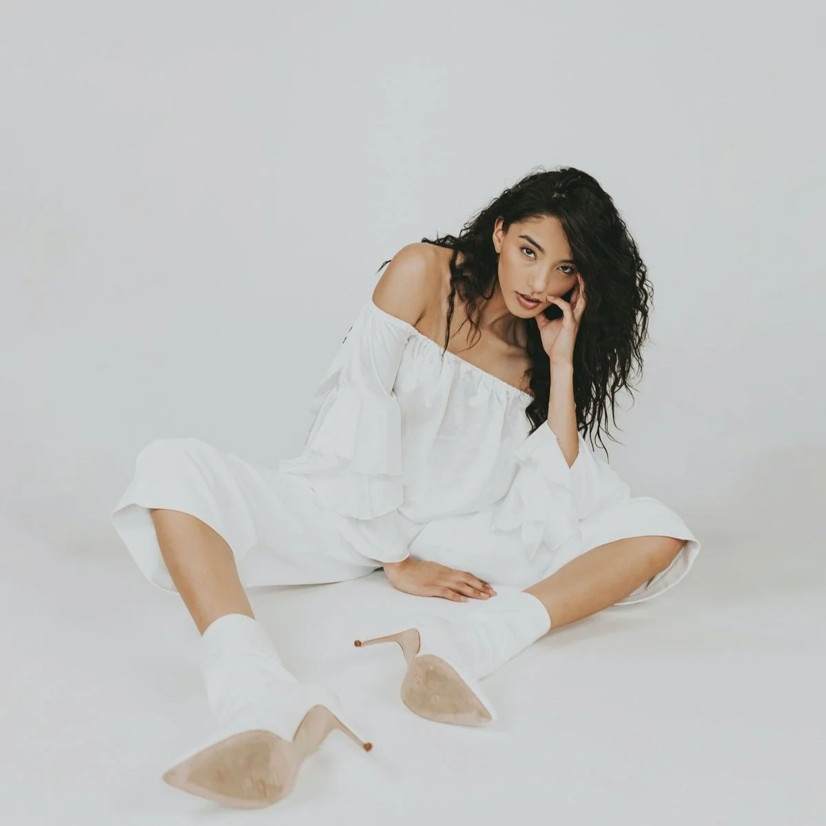A woman with long, curly black hair wearing a white off-the-shoulder dress and beige high heels, sitting on the floor against a plain white background, looking at the camera with her hand near her face.
