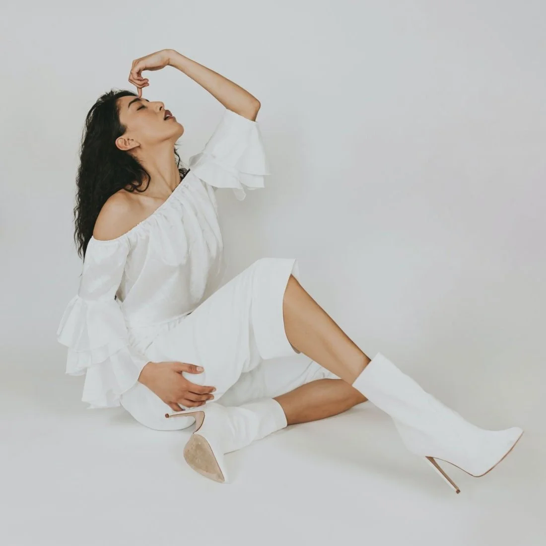 A woman wearing a white off-the-shoulder dress and white high-heeled boots sitting on the floor with one leg extended, her head tilted back, and her eyes closed against a plain white background.