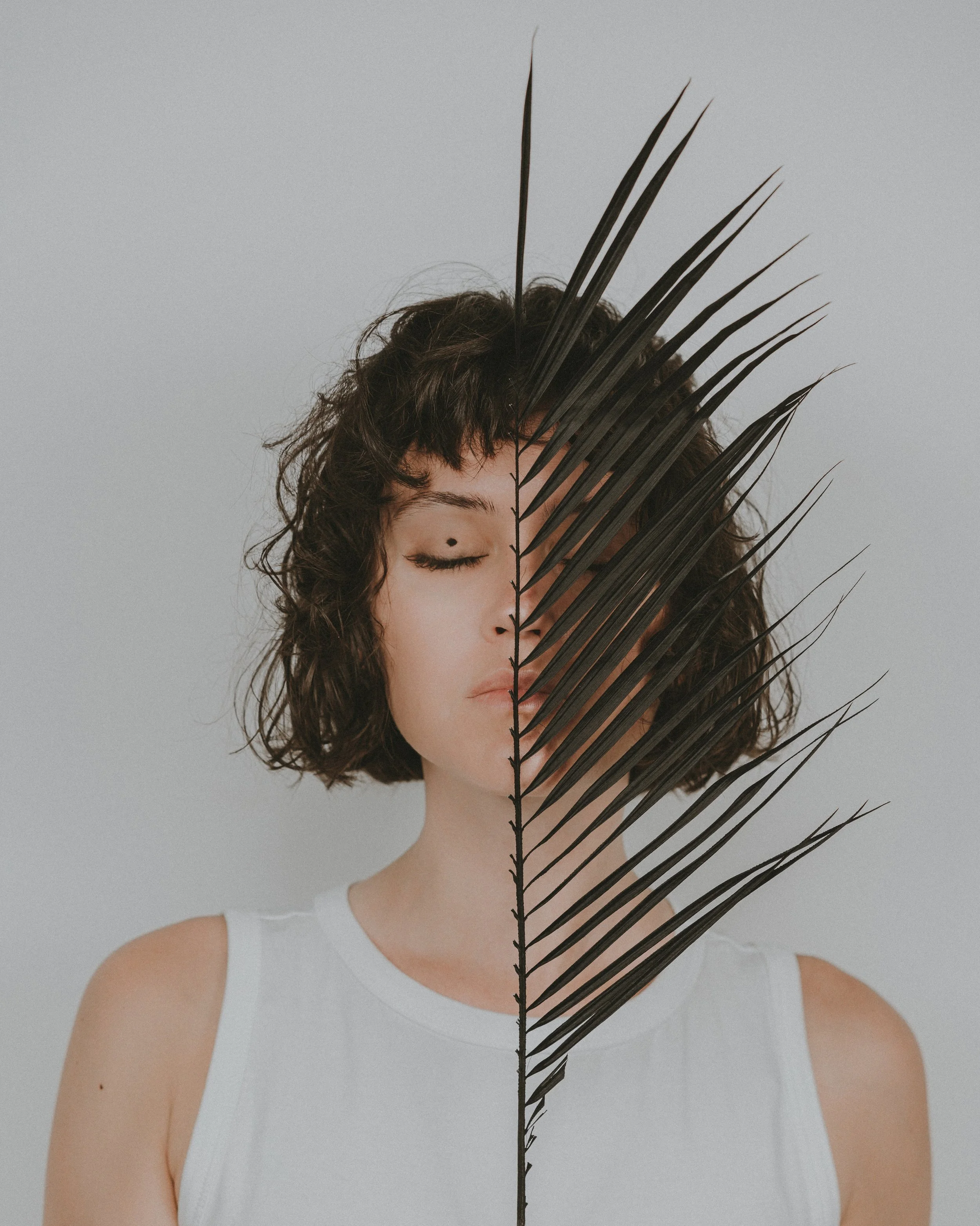 A woman with short curly hair, wearing a white top, has her eyes closed and a black palm leaf covering the right half of her face, against a plain light background.