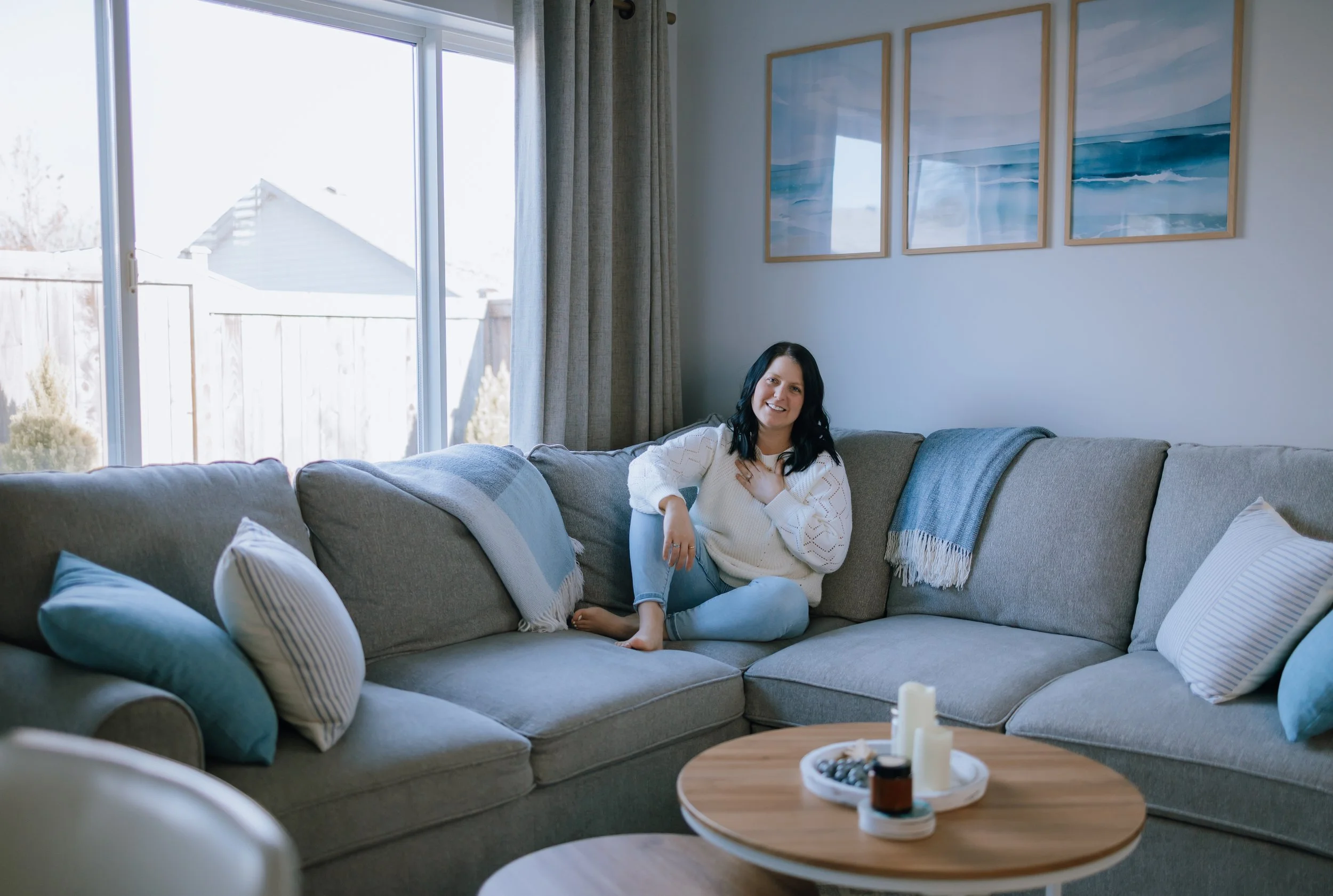 A woman with black hair, wearing a white sweater, sitting on a gray sectional sofa in a living room near a large window with sunlight, outdoor view of a wooden fence, and framed seaside paintings on the wall.