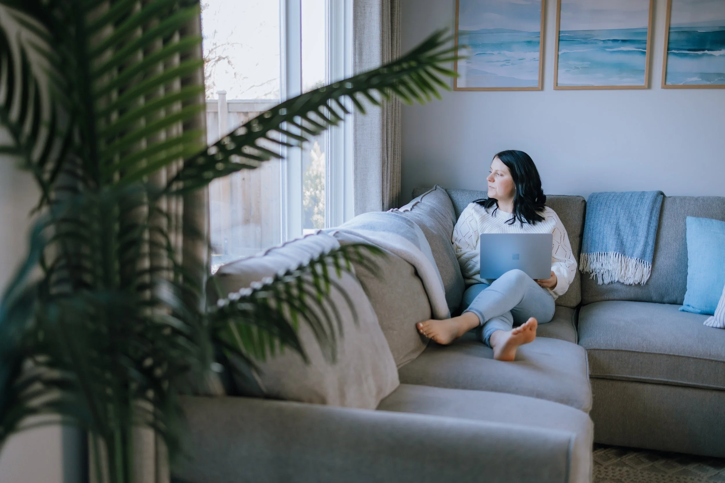 A woman sitting on a gray sofa in a living room, holding a laptop and looking out the window, with blue and white decor, and a large plant in the foreground.
