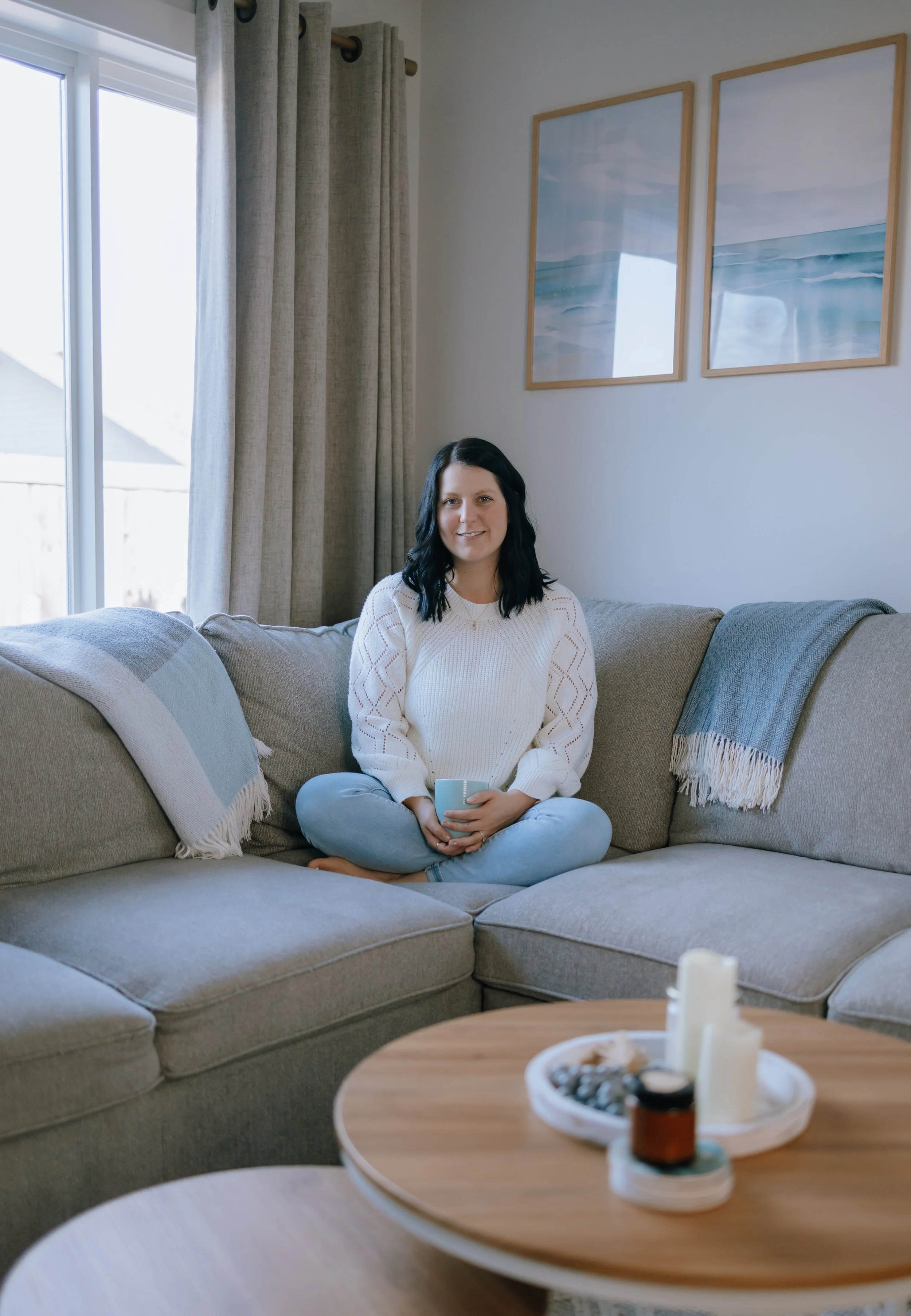 A woman with black hair, wearing a white sweater and jeans, sits cross-legged on a beige sectional sofa in a living room, holding a mug and smiling at the camera. There are decorative pillows on the sofa, and a wooden coffee table with candles and a tray of stones or shells in front of her. Behind her, there are two framed ocean paintings and a window with beige curtains.
