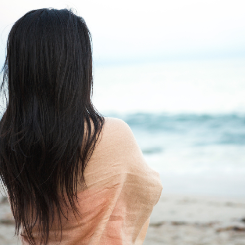 A woman with long dark hair standing on a beach, looking out at the ocean.