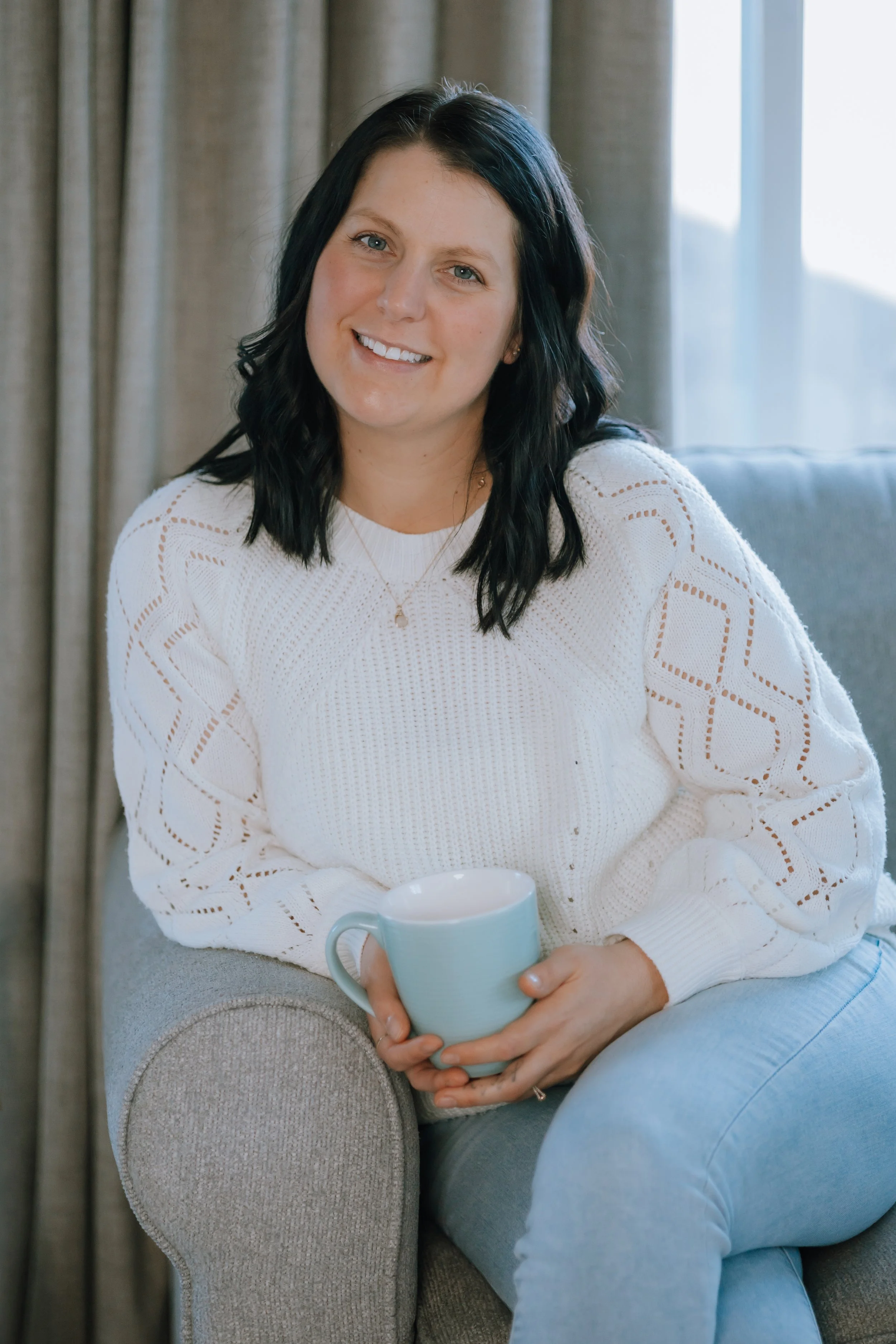 A woman with dark hair wearing a white knitted sweater, sitting on a gray armchair, holding a light blue mug, smiling at the camera, in a cozy indoor setting with curtains and a window.