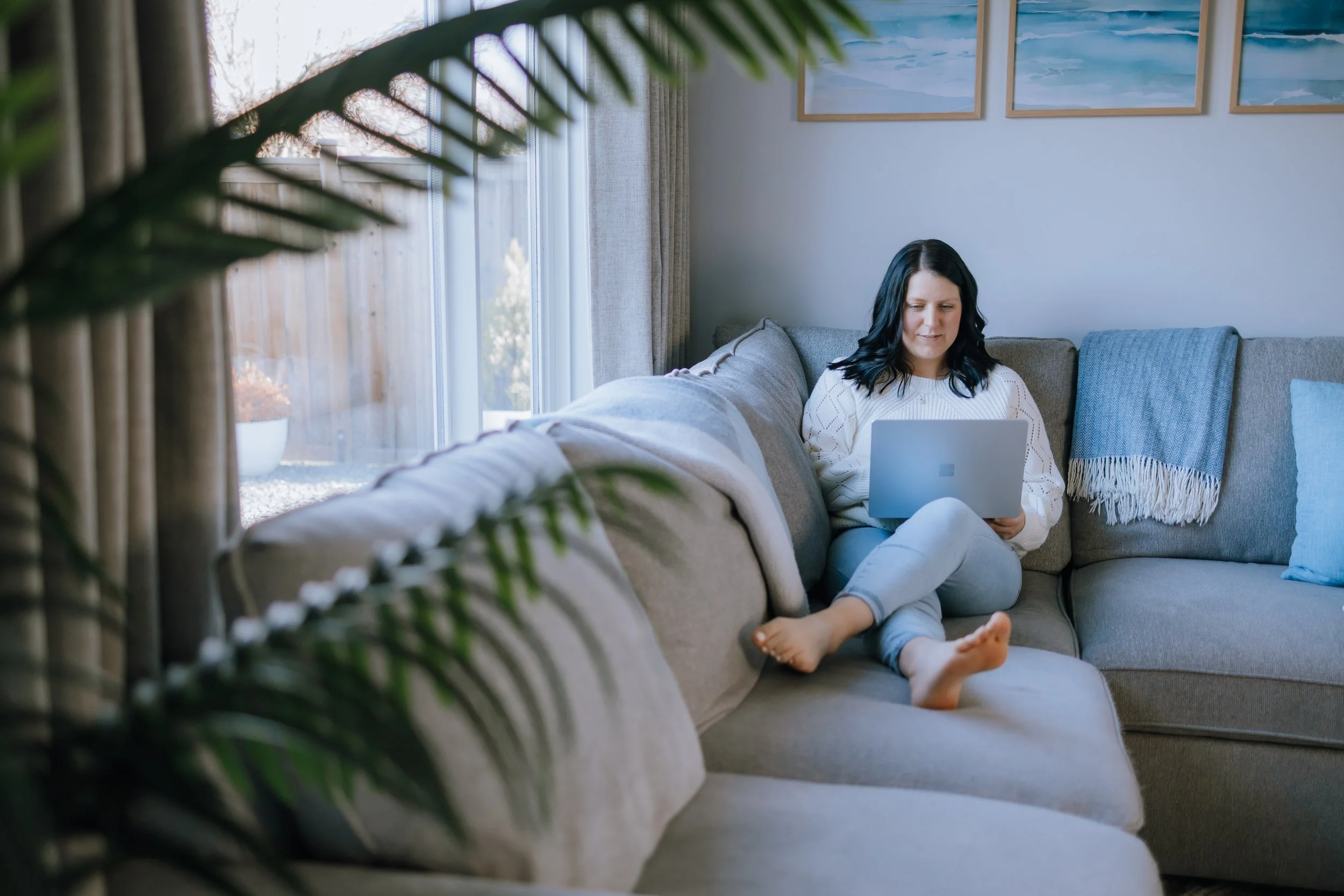 A woman with black hair sitting on a gray sectional sofa, using a silver laptop. She is barefoot, wearing a white sweater and light blue jeans. The room has a large window with curtains, some framed landscape artwork on the wall, and a blue throw blanket on the sofa.
