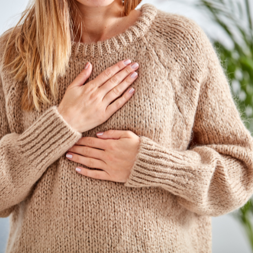 A person wearing a beige sweater with one hand on their chest and the other on their abdomen.