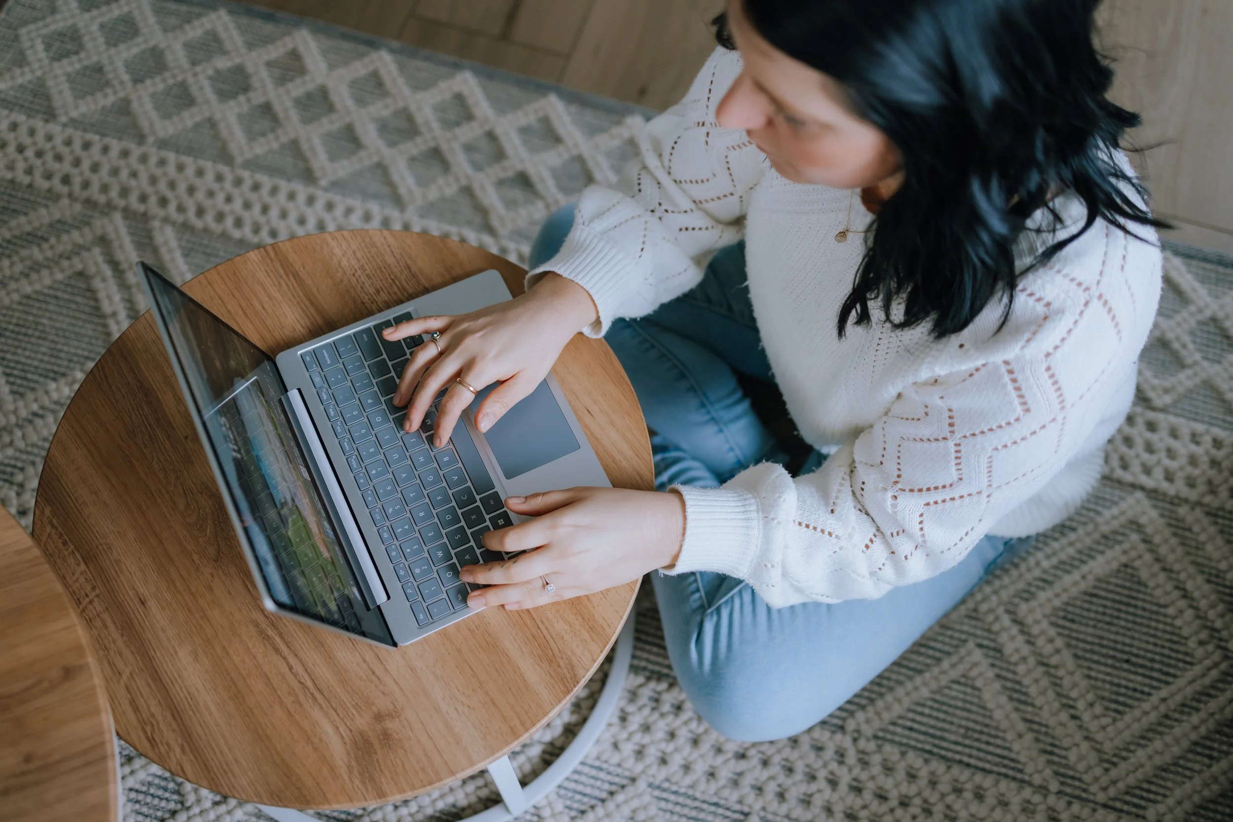 A woman with black, wavy hair wearing a white knitted sweater and light blue jeans using a laptop on a round wooden table, viewed from above.