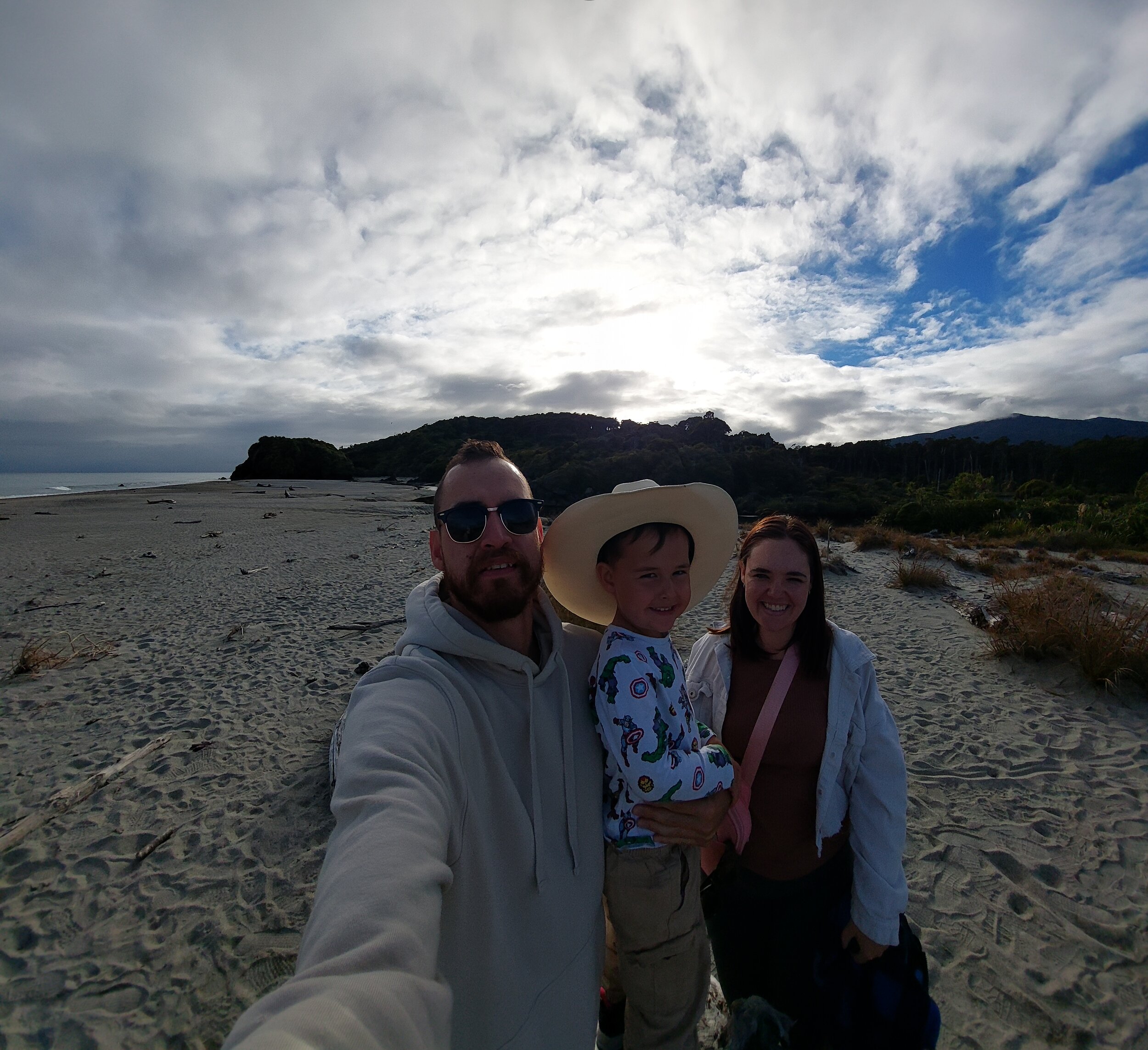 A family of three taking a selfie on a sandy beach with cloudy skies and a mountain in the background.