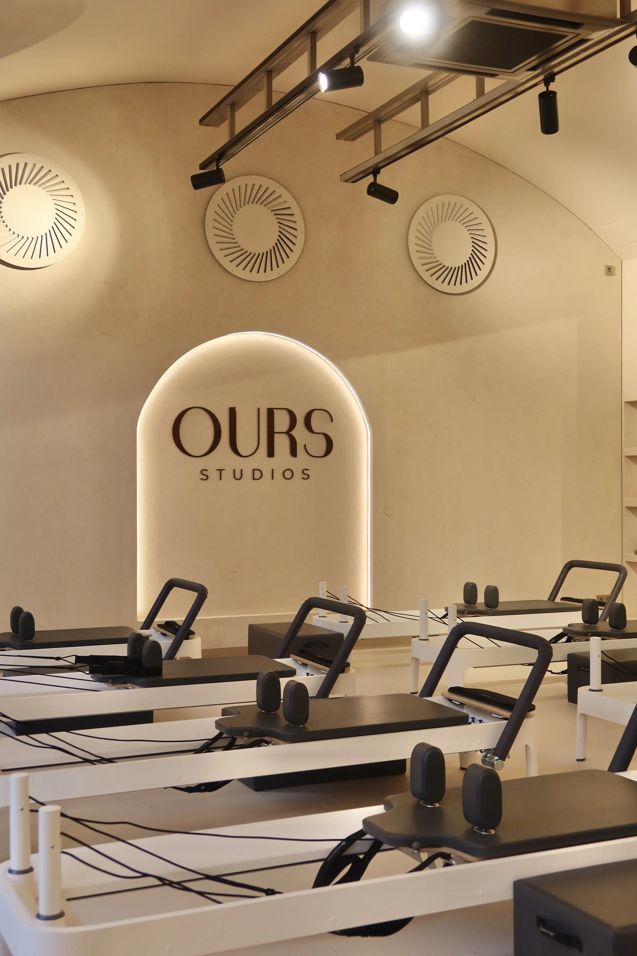 Leerer Pilates- oder Yoga-Raum mitliegendem schwarzen Geräte auf weißen Matten, beleuchtetem Firmenlogo 'OURS STUDIOS' an der Wand, runden Lüftungsgittern an der Wand und Deckenleuchte in der Decke.