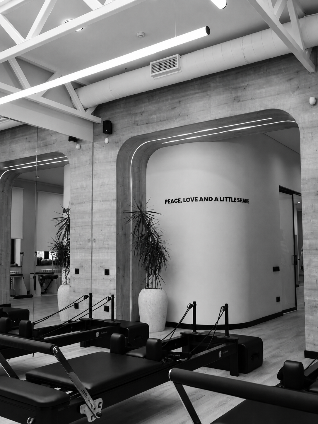 Inside a modern fitness studio with Pilates reformers, large potted plants, a mirror wall, and a sign reading 'Peace, Love and a Little Shake' on the wall.