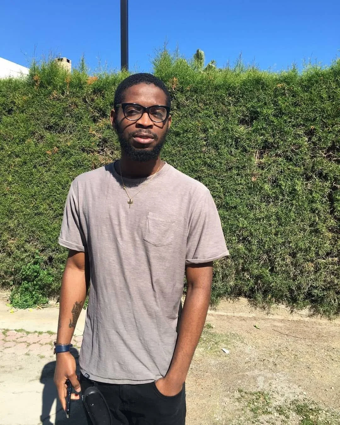A man resting his chin on his hand, looking into the camera. He has short hair, a beard, and wears a white shirt. The background is a plain wall with a blurred poster or chart.