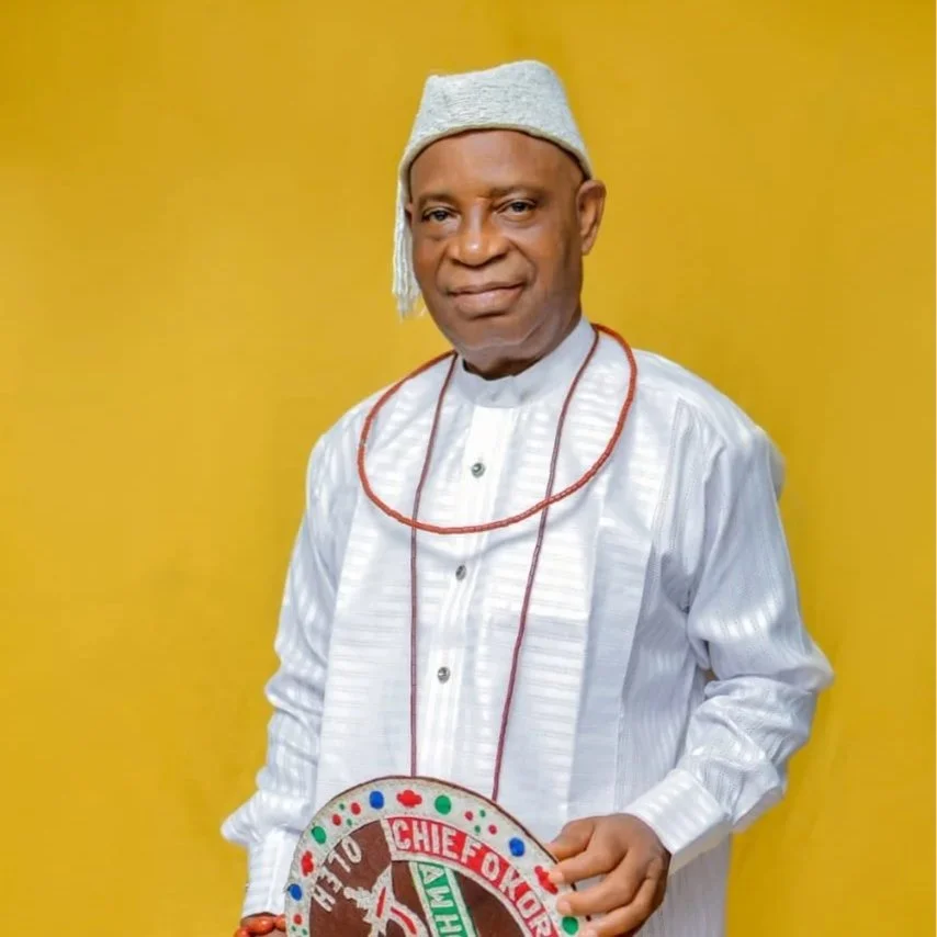A man dressed in traditional Nigerian attire, wearing a white agbada, a gray cap, and holding a colorful beaded shield with Nigerian symbols and the words "Chief of Staff" against a yellow background.