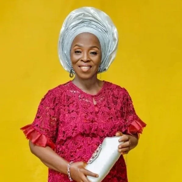A woman in a pink lace dress and silver gele headwrap holding a white clutch purse, standing against a yellow background.