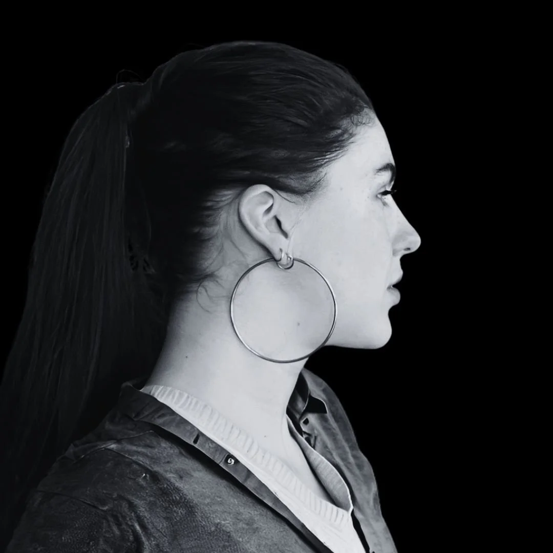 Side profile of a woman with long dark hair tied back, wearing a large hoop earring and a dark jacket, against a black background.