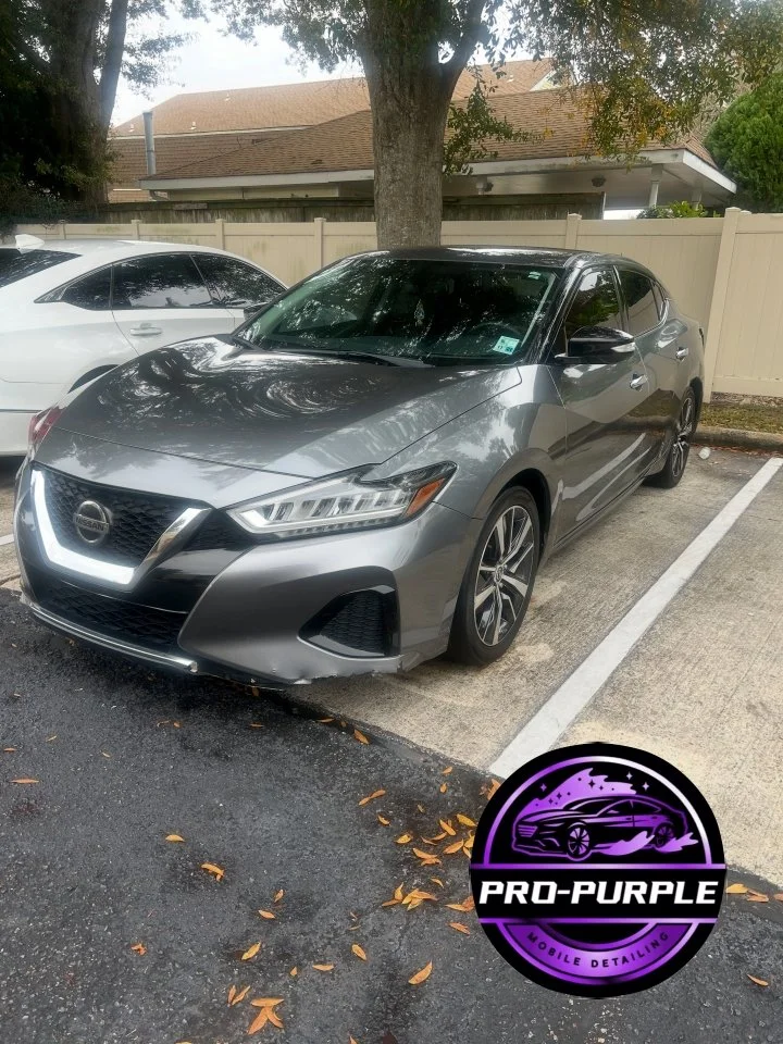 A silver Nissan sedan parked in a parking lot near a tree with a white fence in the background. There is a purple and black logo in the bottom right corner that says 'PRO-PURPLE' with a stylized car and the text 'MOBILE DETAILING'.