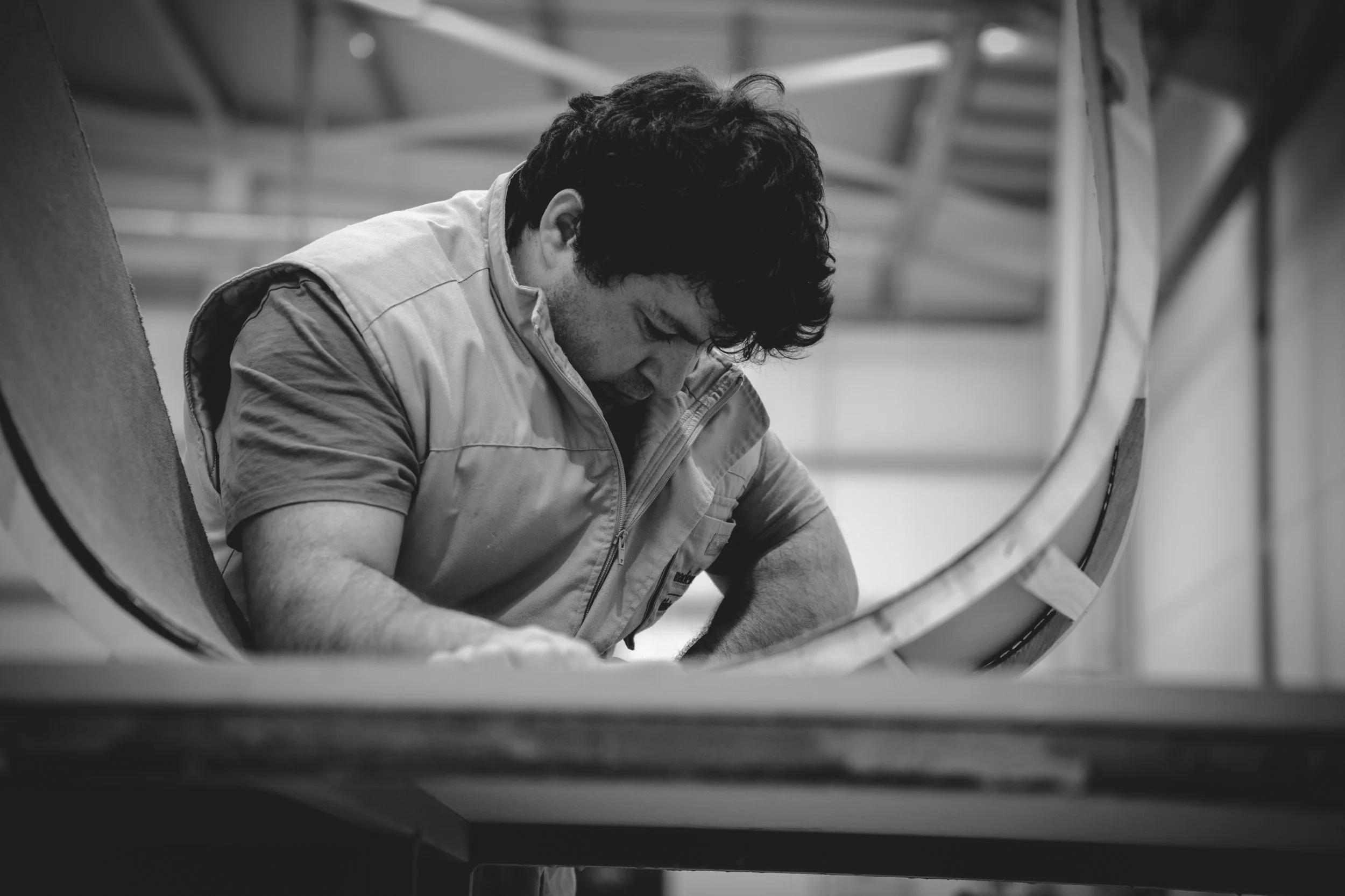 Black and white photo of a man working inside a large circular metal structure, with his head bent down and wearing a short-sleeved shirt and a vest.