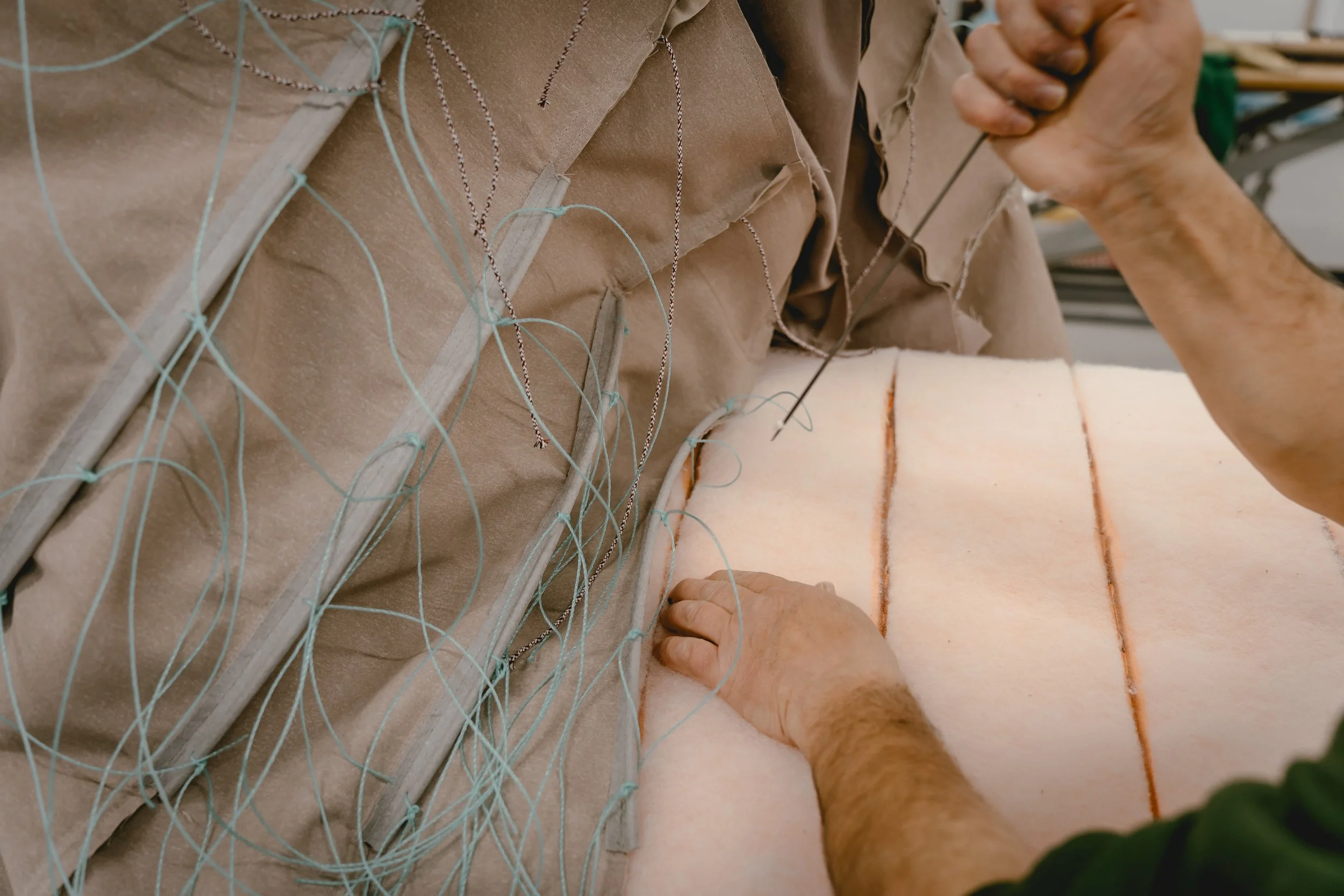 Person sewing the edges of a thick, cushioned fabric with a needle and thread on a worktable.