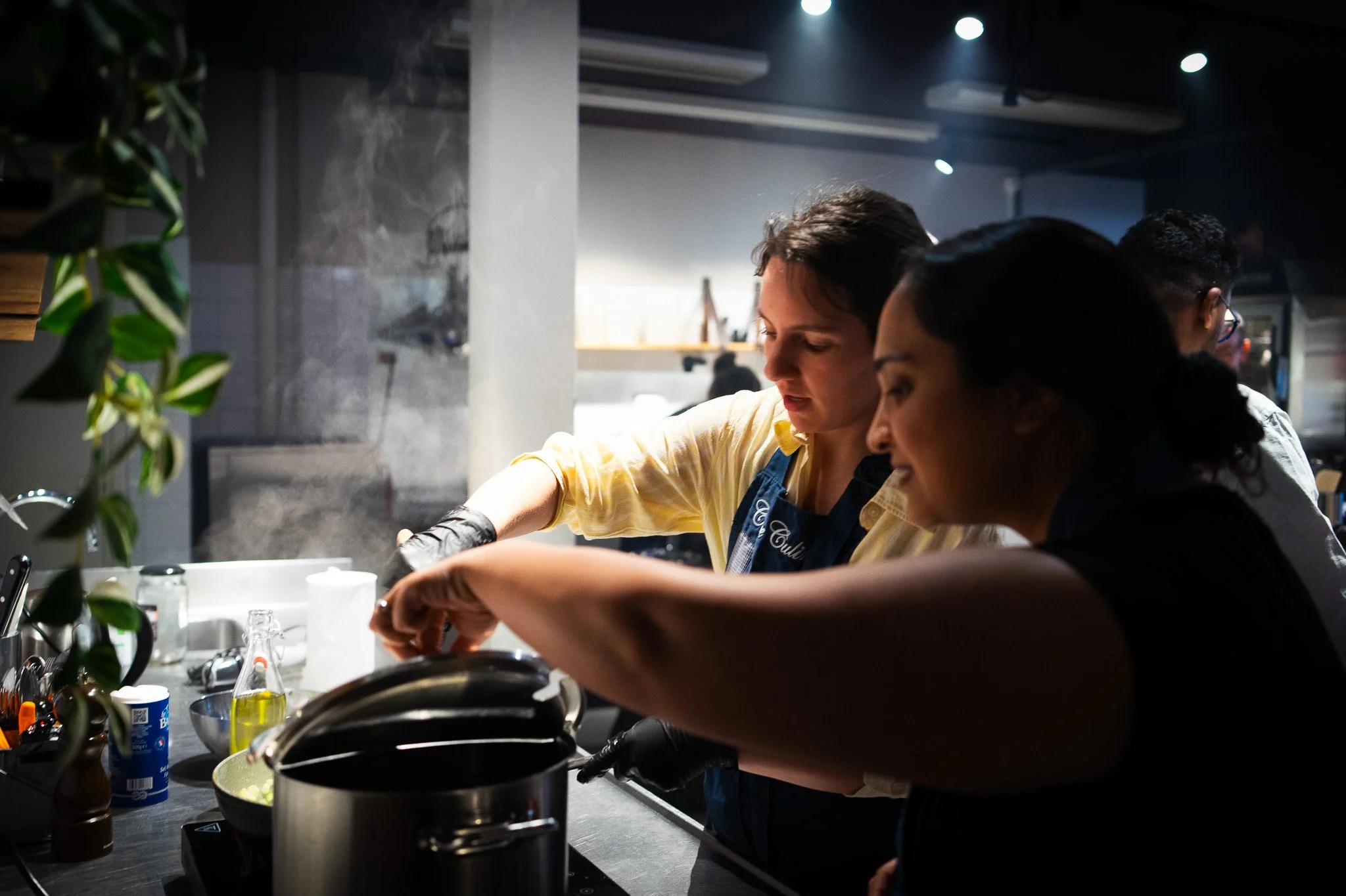 People cooking in a modern kitchen with overhead lights, with various utensils and ingredients on the counter, including a bowl, a can, and oil, and a leafy plant on the left.