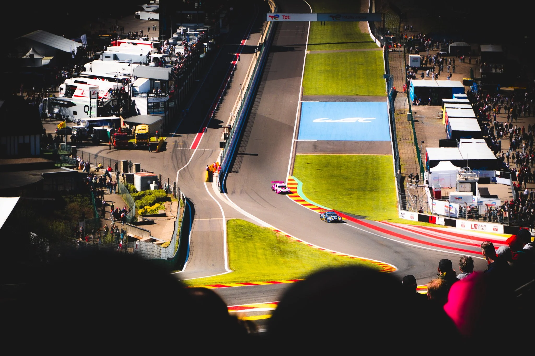 An aerial view of a race track during a motorsport event, featuring racing cars on a curved track, spectators in the stands, and team trailers in the paddock area.