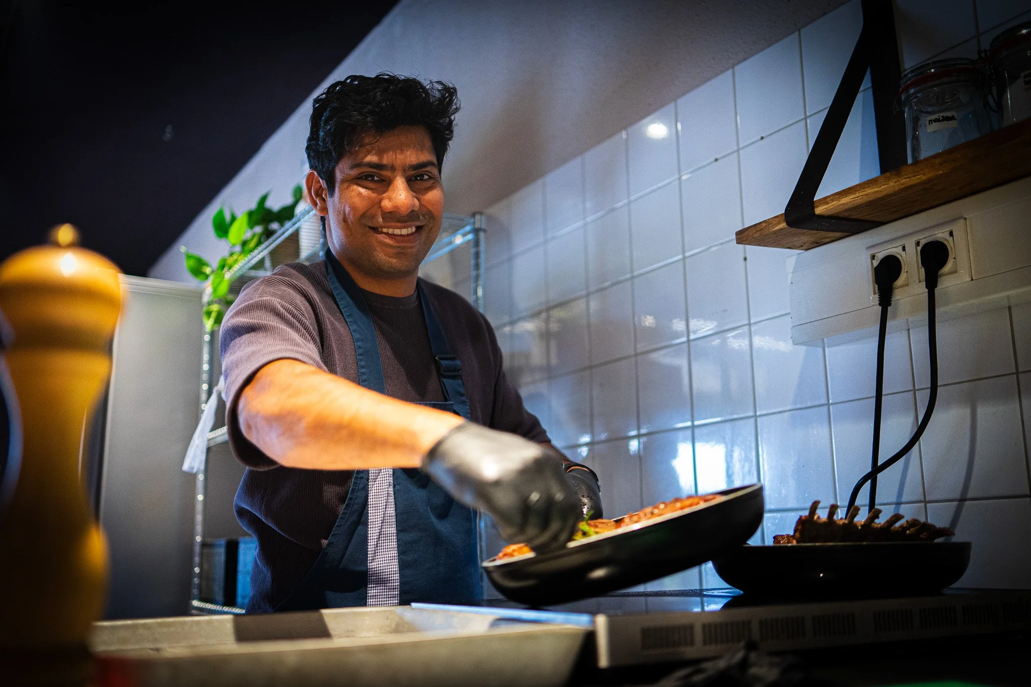 A man wearing an apron and gloves cooking with a smile in a modern kitchen, with a tiled white backsplash and a shelf with jars, in front of a skillet on a stove.