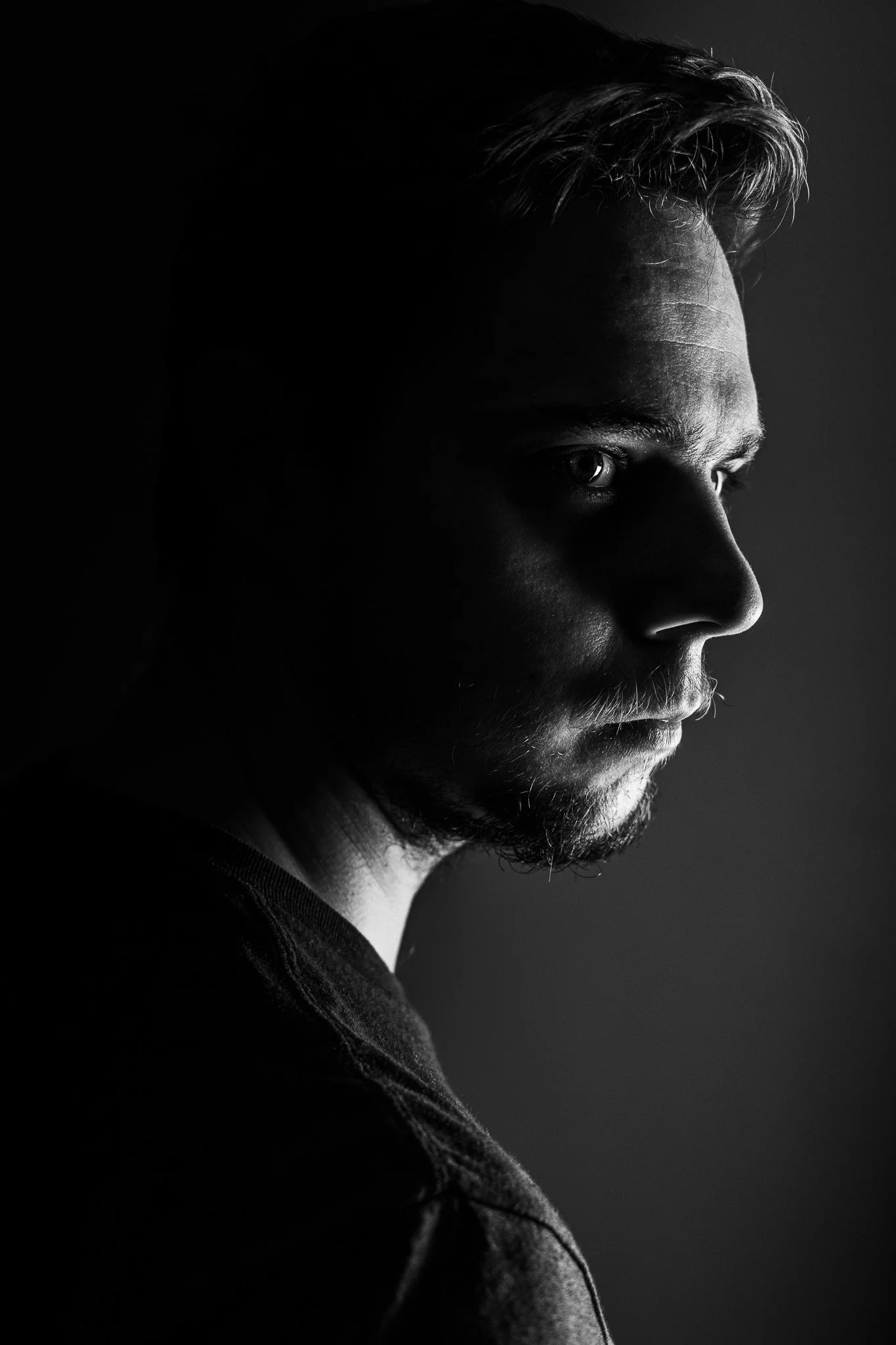 Black and white portrait of a man with light illuminating one side of his face, showing short hair, beard, and a serious expression.
