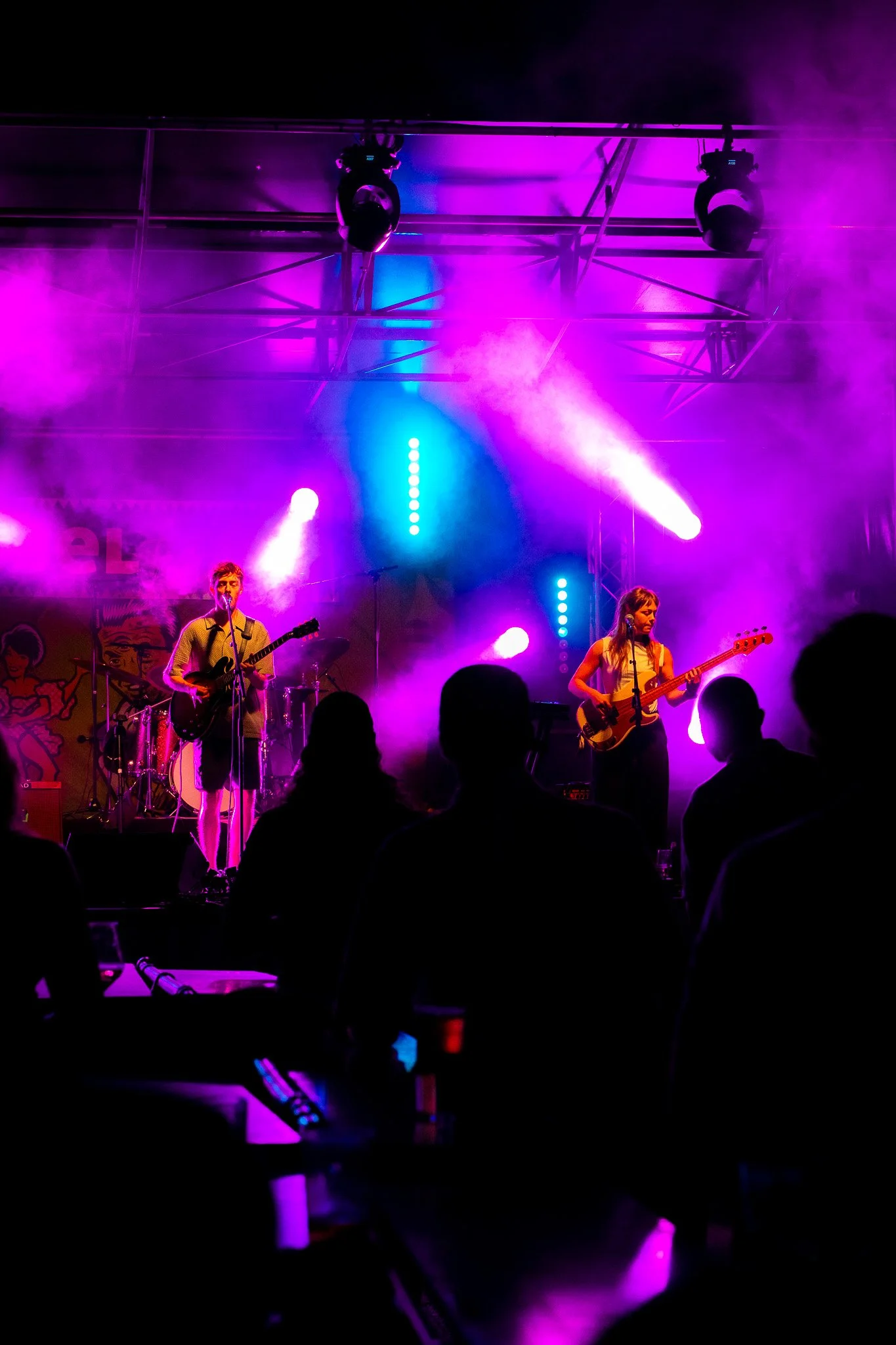 A band performs on stage under purple and blue lights with fog effects, with audience silhouettes in the foreground.
